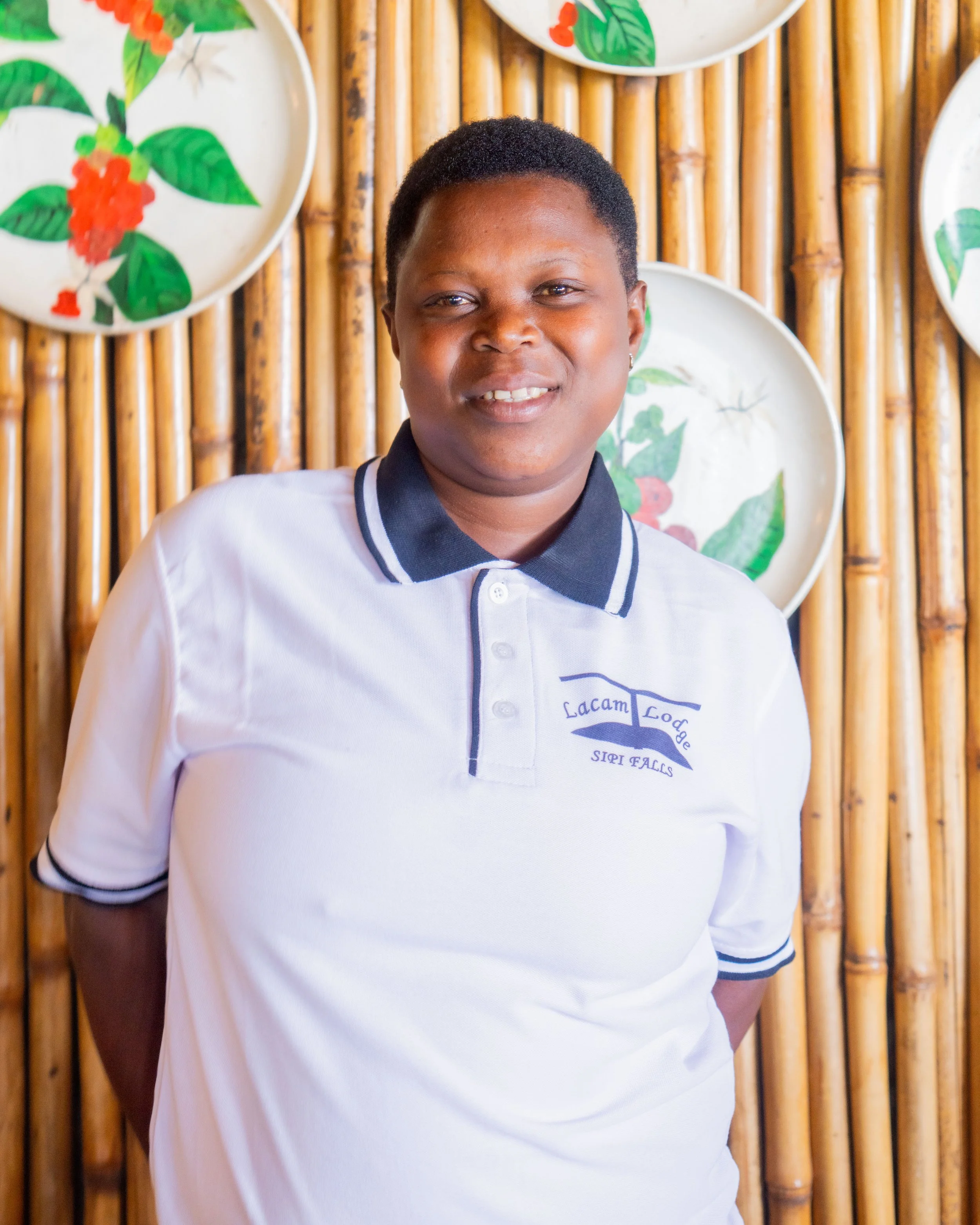 A woman with short hair, wearing a white polo shirt with dark collar and sleeve trims, standing in front of a bamboo wall with decorative plates featuring green and red tropical foliage.