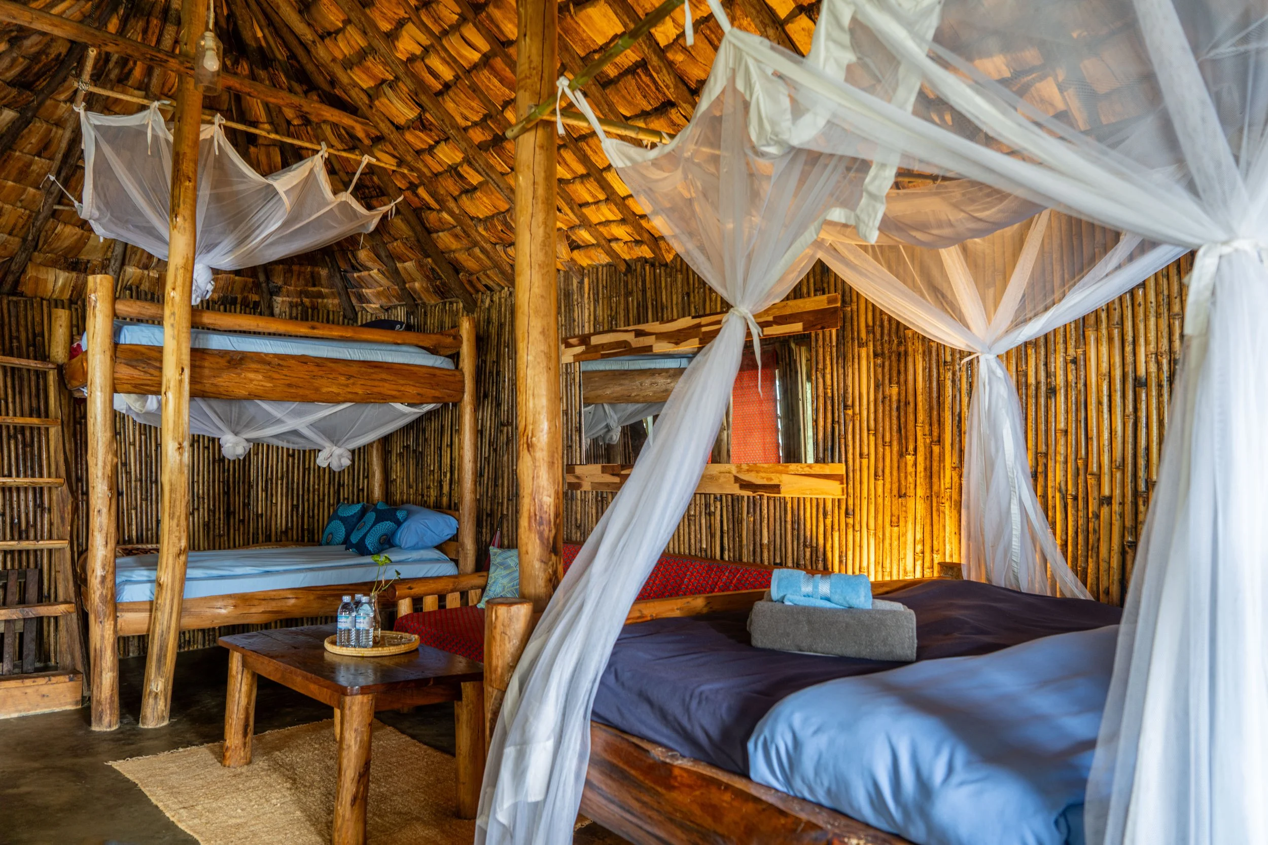 Bedroom with wooden furniture and bamboo walls, featuring a large wooden bed with a mosquito net, a bunk bed, and a small table with water bottles.