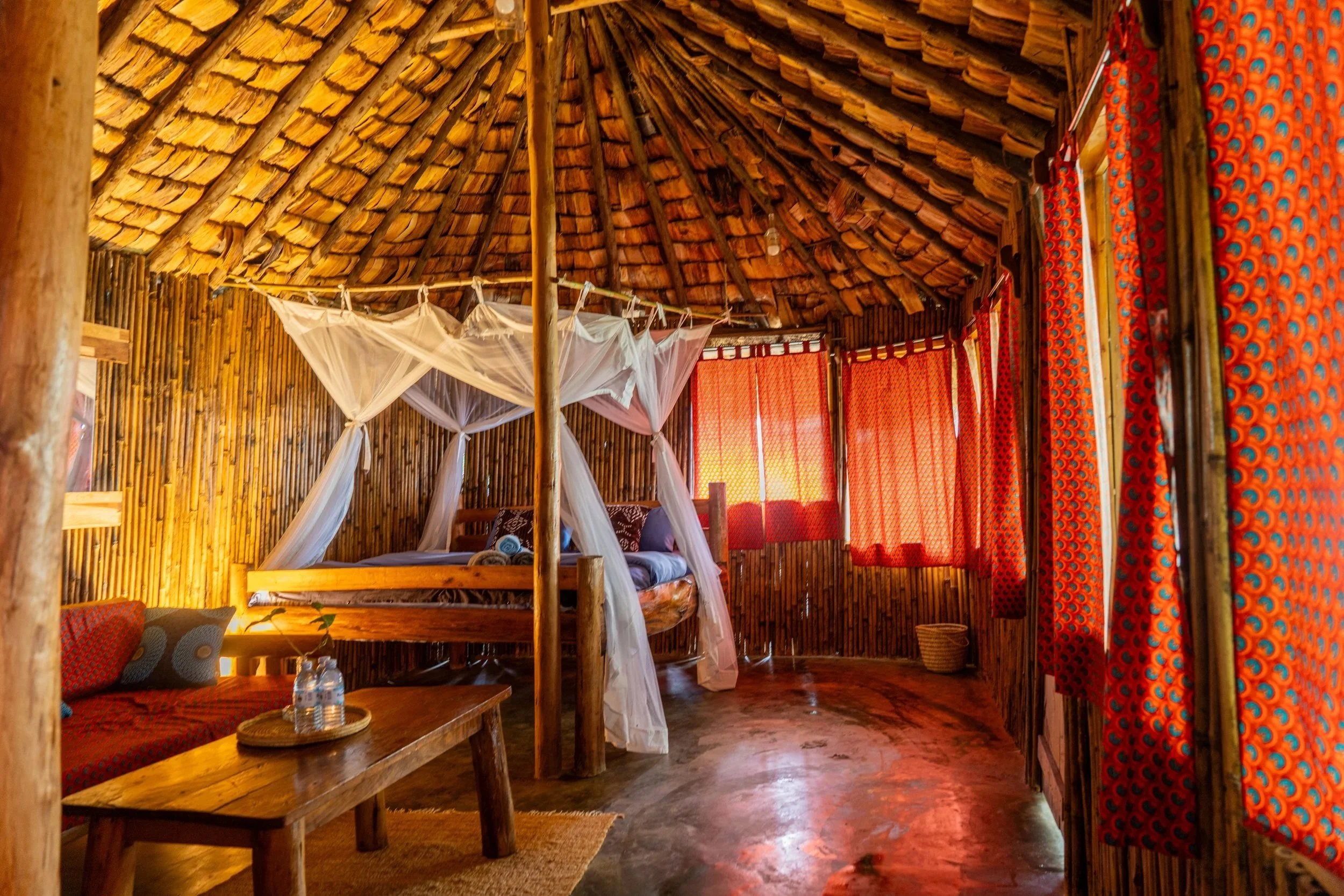 Interior of a rustic bamboo hut with a four-poster bed with white mosquito netting, red patterned curtains, a small wooden table with bottled water, a cushioned seating area, and a woven basket in the corner.