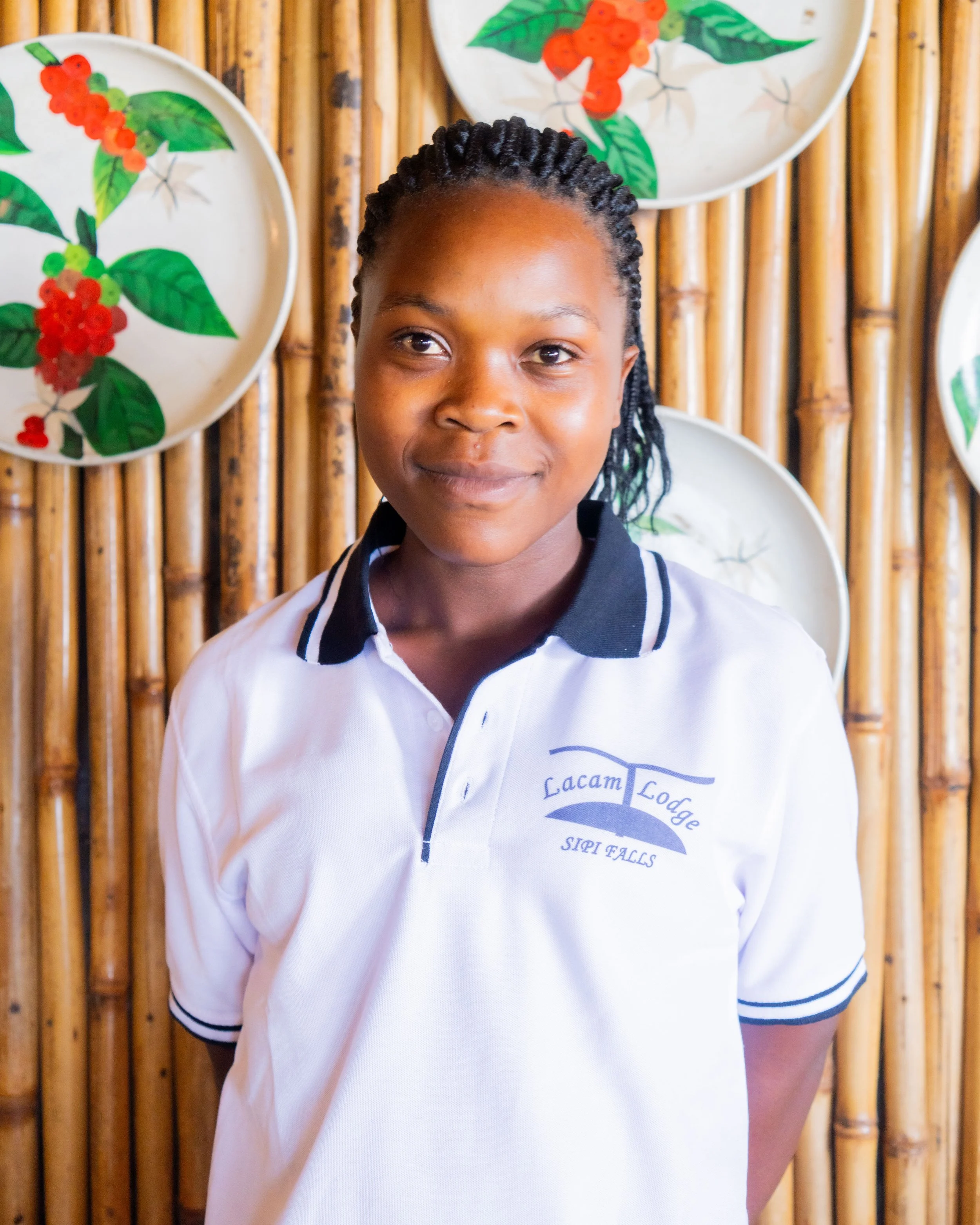 A young woman in a white polo shirt with black collar and trim, standing in front of a bamboo wall decorated with patterned plates featuring red and green leaf designs.
