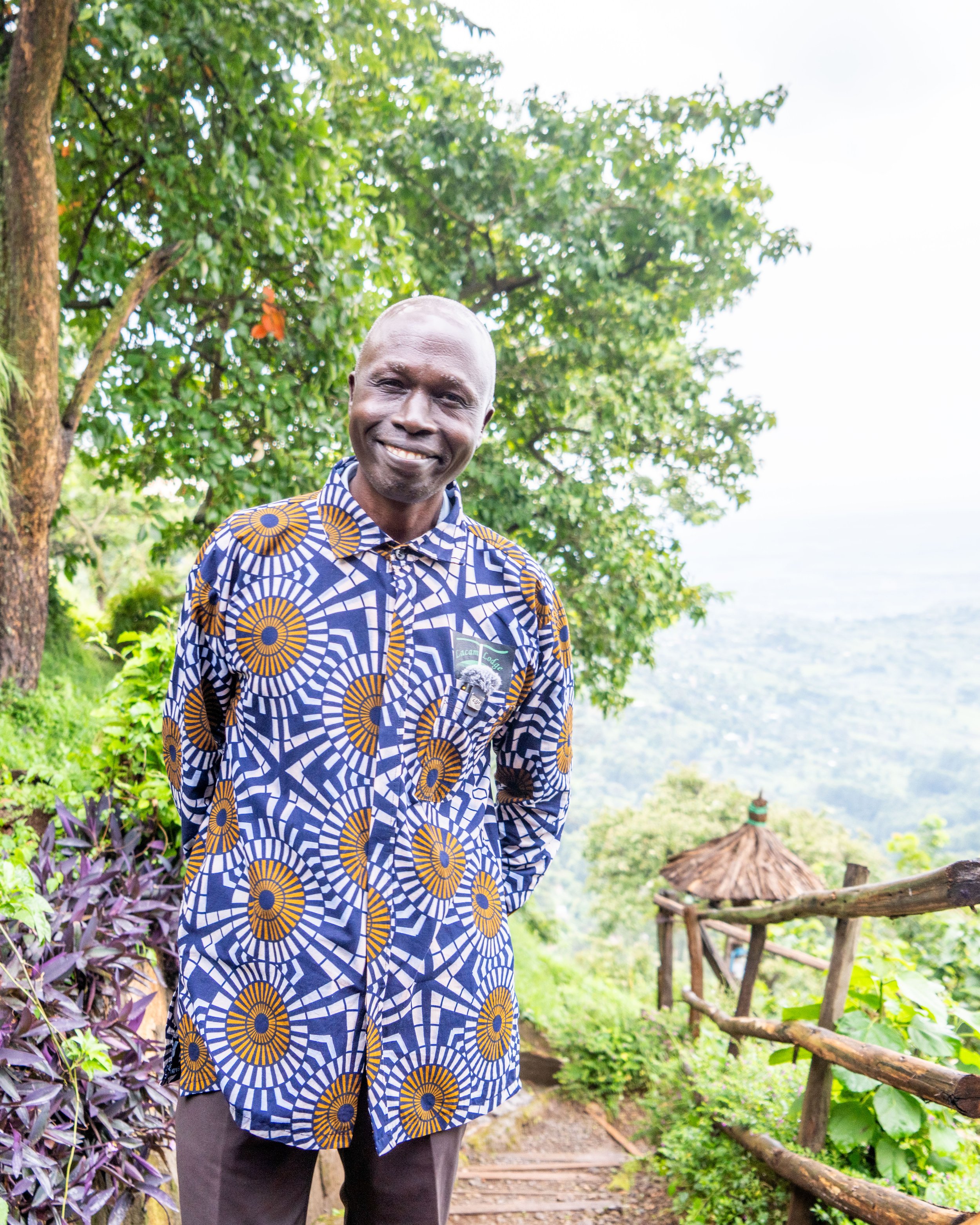 A smiling man in a patterned shirt standing outdoors on a trail overlooking a lush landscape with trees and a distant view.