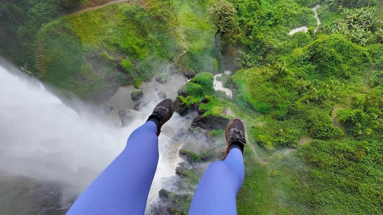 Person standing on a ledge high above a lush green forest and waterfall, with steep cliffs below.