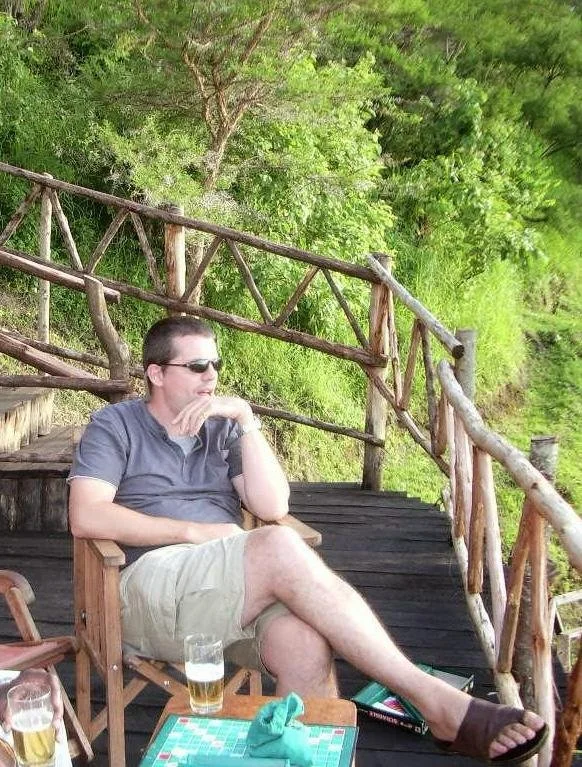 Man sitting outdoors on a wooden deck with a glass of beer, wearing sunglasses, gray shirt, and shorts, with a green lush background and rustic wooden railing.