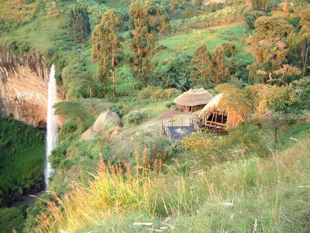 A lush green landscape with a waterfall on the left side and trees surrounding a small rustic hut on a hillside.