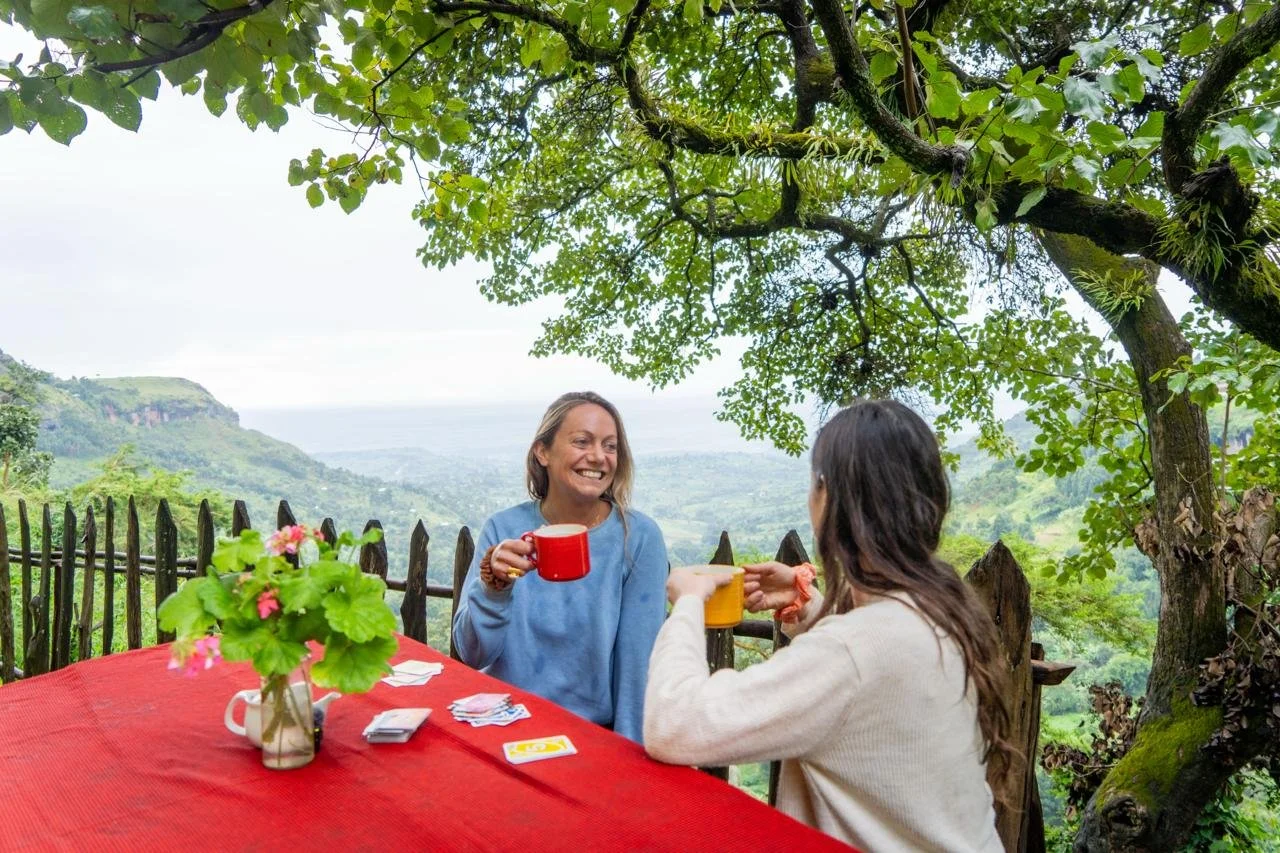 Two women sitting at a red table outdoors under a large tree, playing cards and holding mugs, with a scenic view of green hills and a cloudy sky in the background.