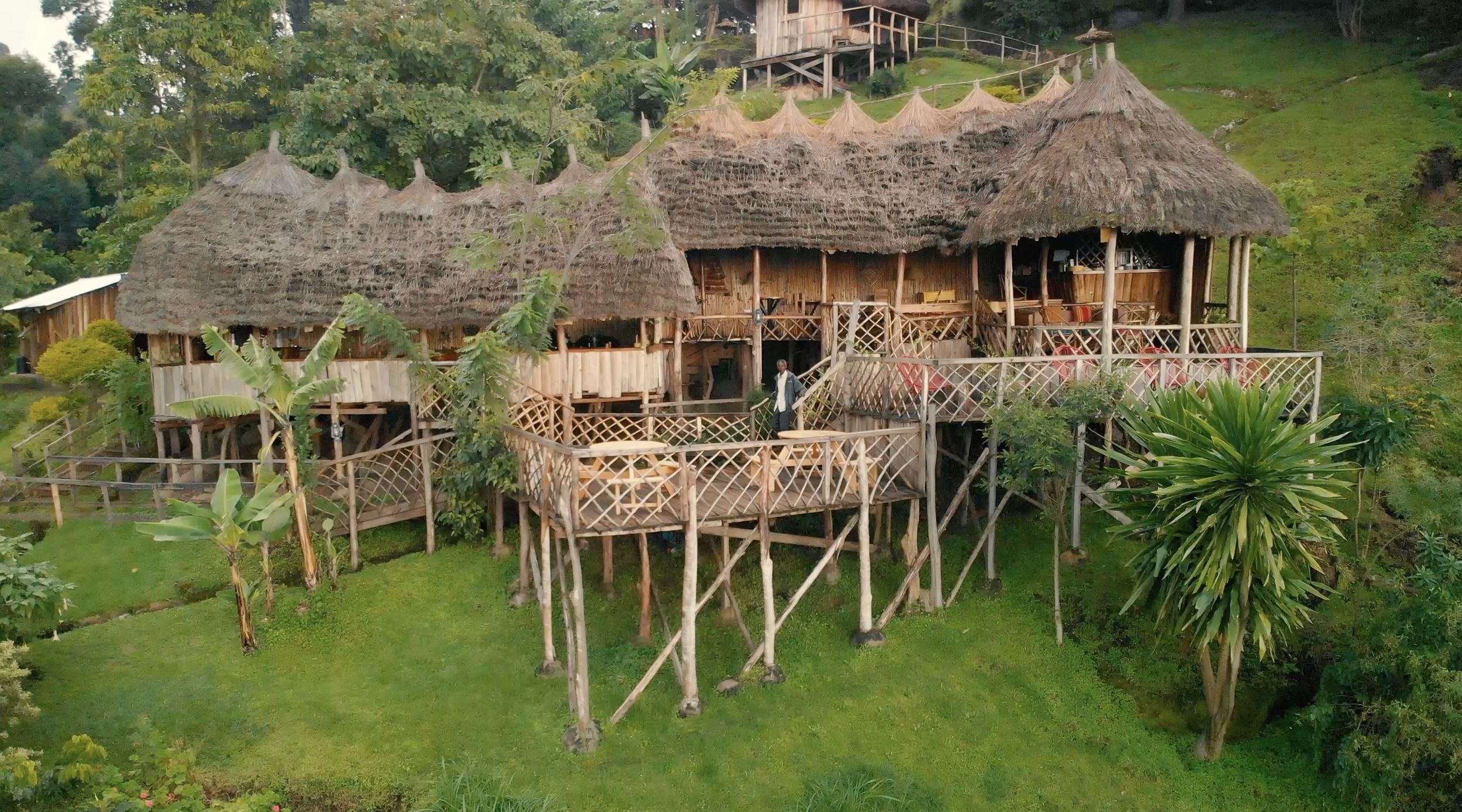 A large elevated wooden house with thatched roof, situated in a lush green tropical landscape with trees and grass. There is a man standing on the deck, and other buildings can be seen in the background.