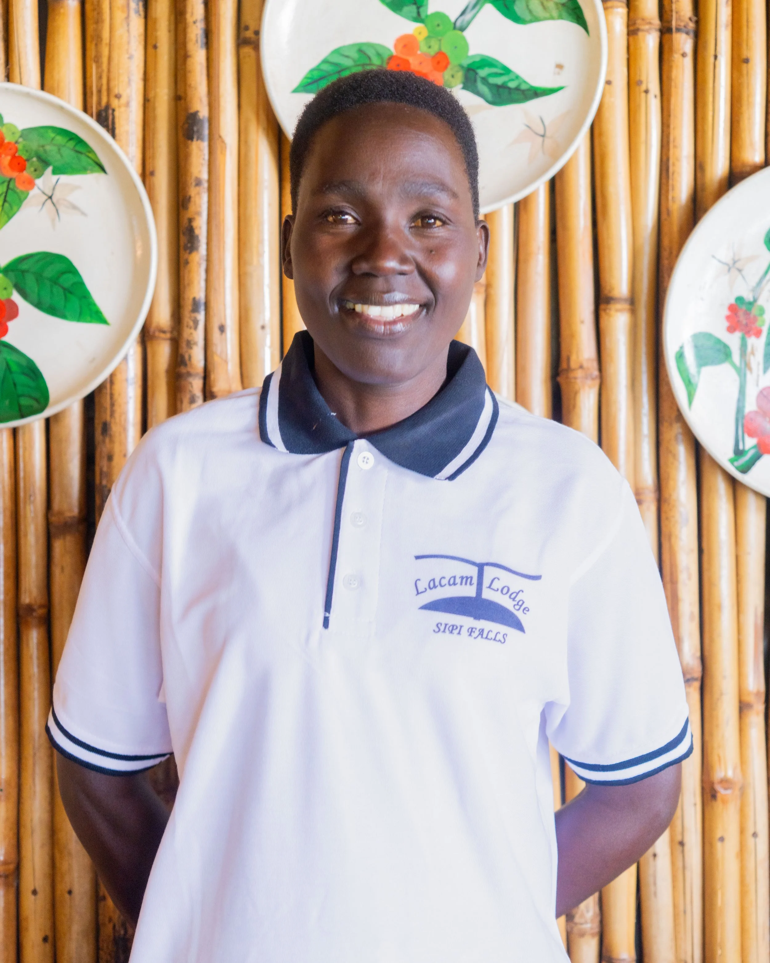 A smiling person standing in front of a bamboo wall decorated with painted plates featuring green leaves and red berries, wearing a white polo shirt with a dark collar and logo.