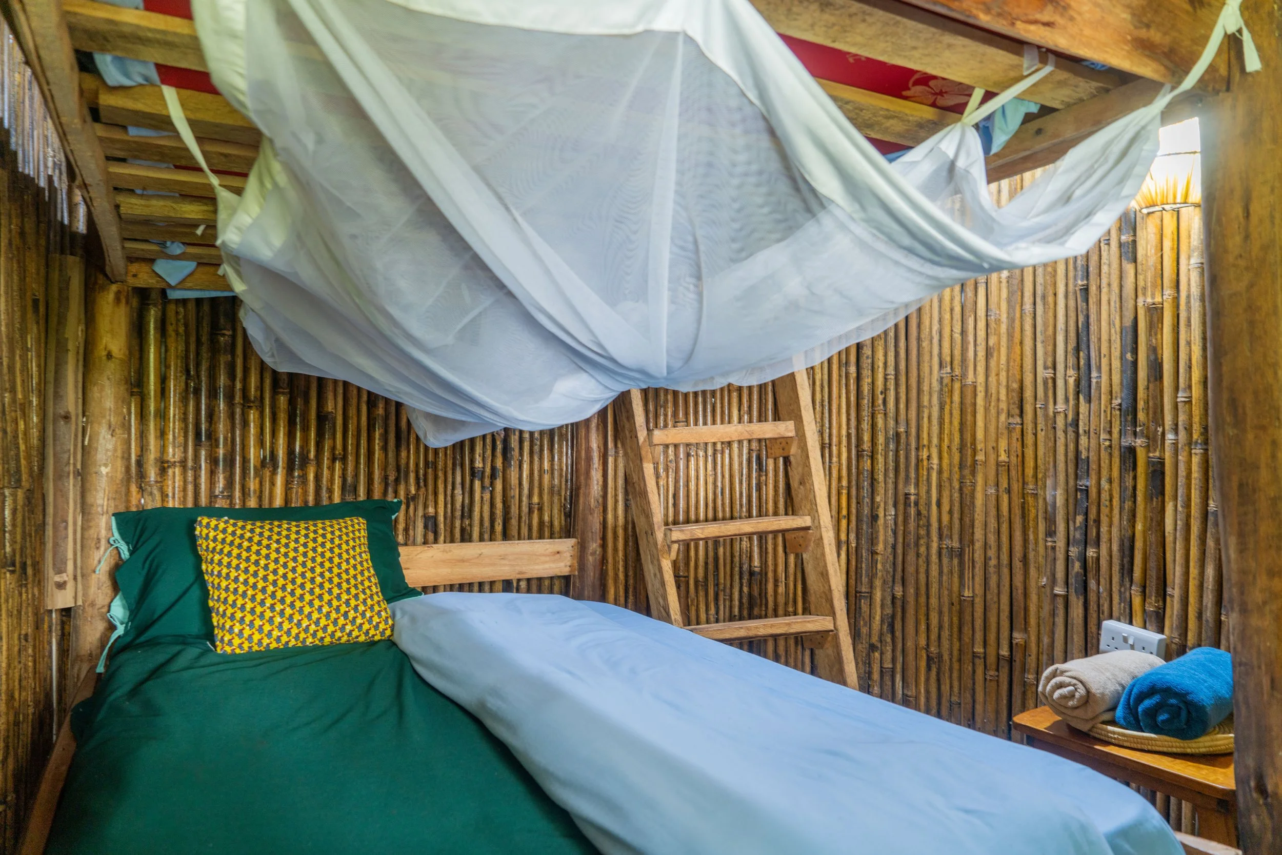 A rustic bedroom with bamboo walls, a wooden bed with green bedding, a yellow patterned pillow, a ladder, and a mosquito net hanging from the ceiling. There are rolled towels on a small table near the electrical outlet.