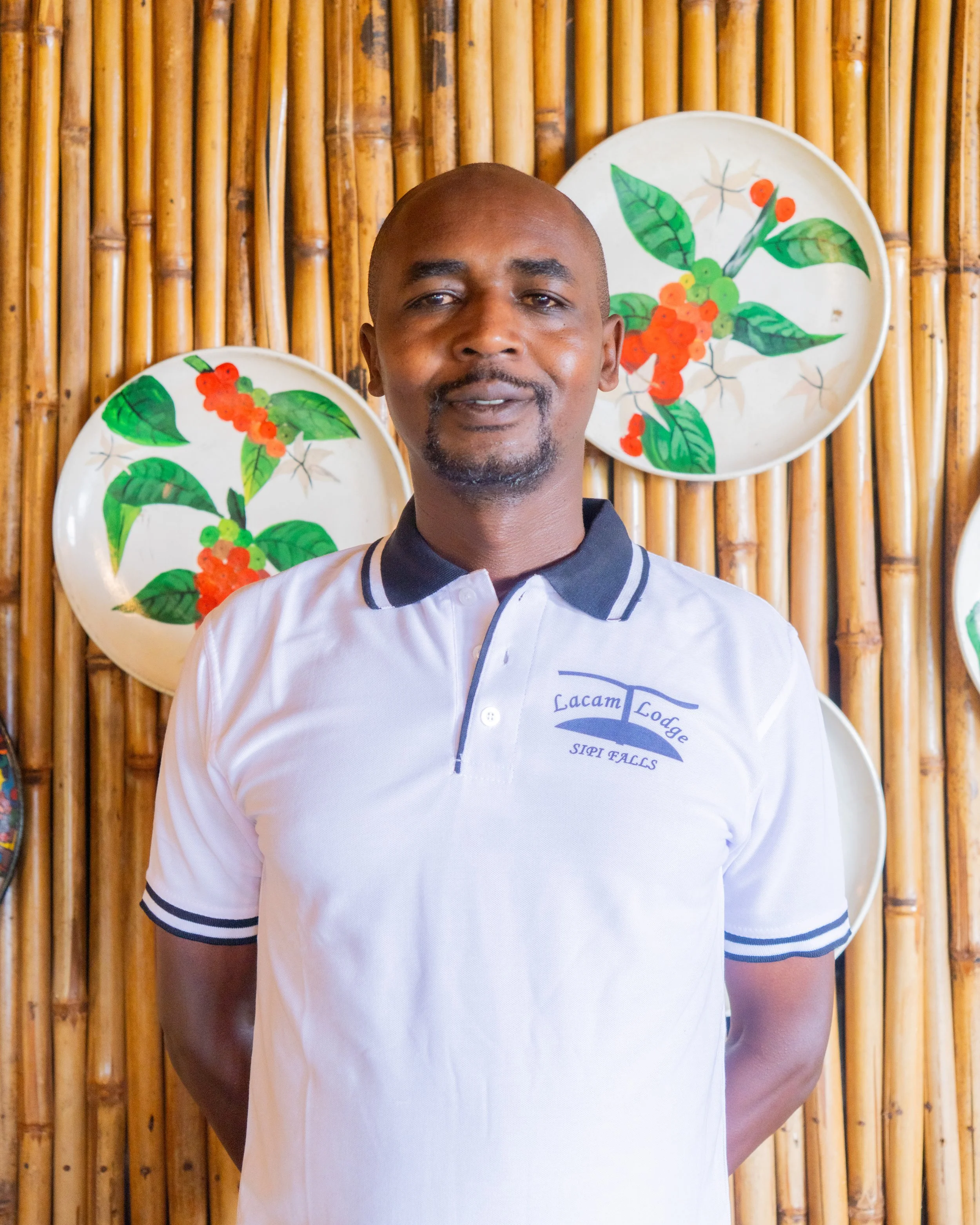 A man standing in front of a bamboo wall with decorative plates featuring green leaves and orange-red berries, wearing a white polo shirt with black collar and sleeve accents, and text that reads 'Lacam Lodge SIPI FALLS.'