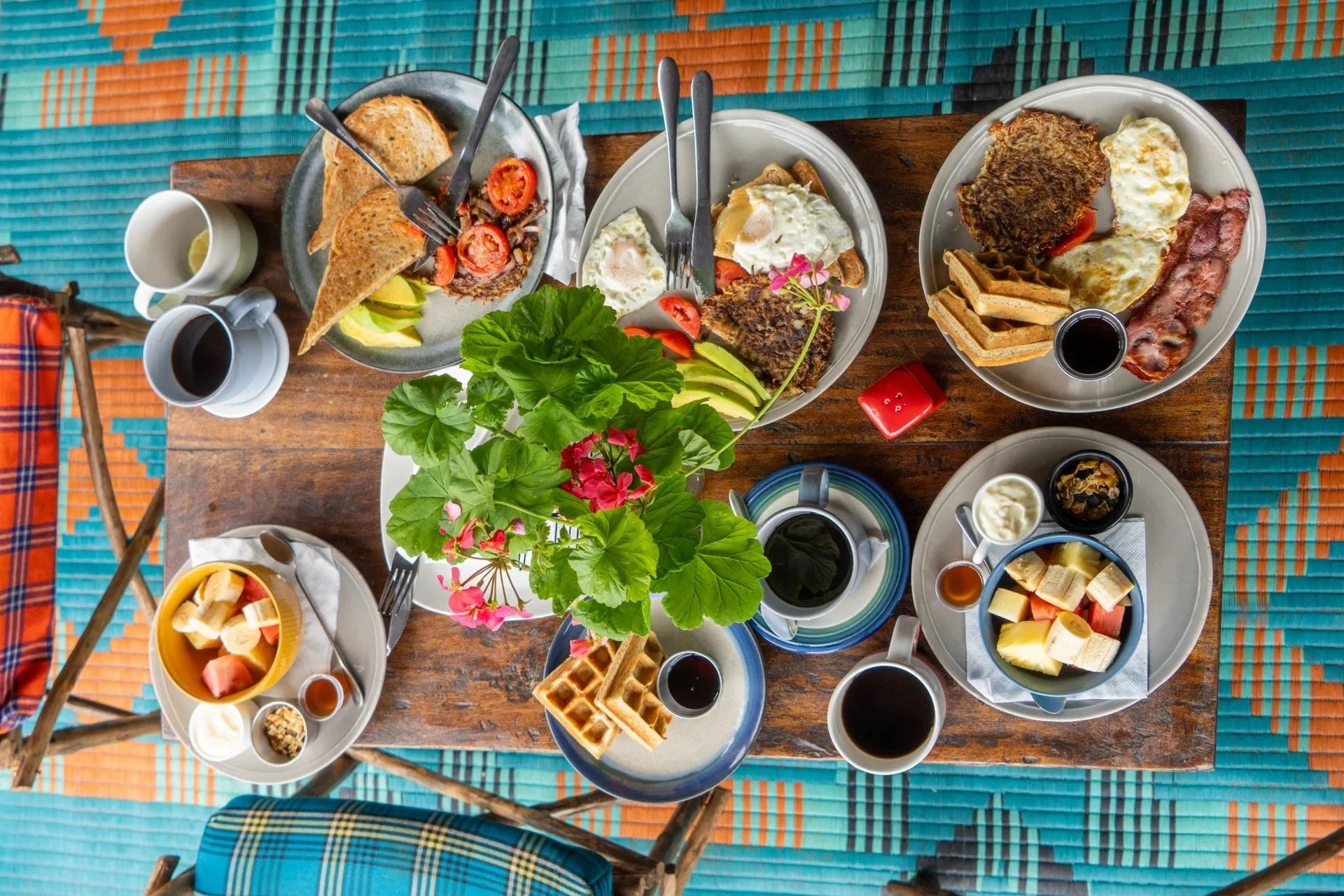 A top-down view of a wooden table set with various breakfast foods, beverages, and a plant centerpiece, with a colorful woven mat underneath.