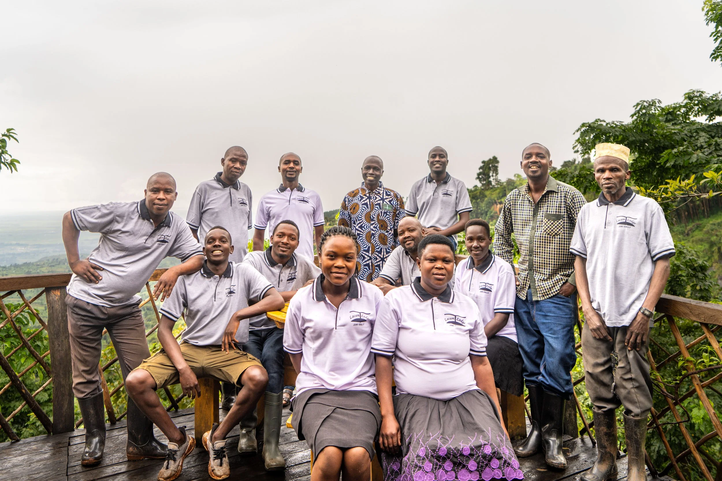Group of fourteen people outdoors on a wooden deck with a scenic green landscape in the background. The group includes men and women, some wearing matching gray polo shirts and others in patterned or plain clothing, posing for a photo.