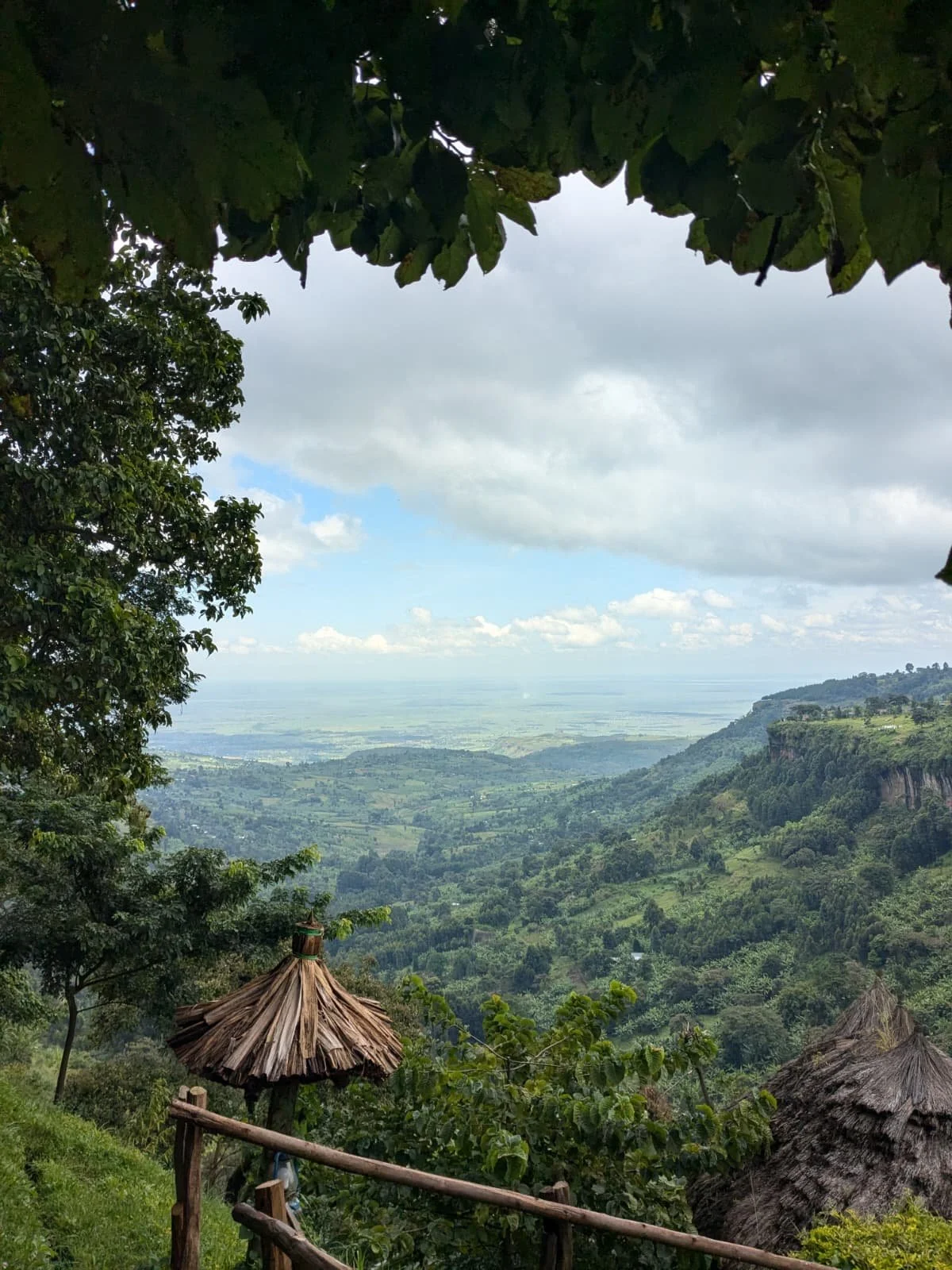 A scenic view of a lush green valley with hills under a partly cloudy sky, framed by trees and thatched-roof huts in the foreground.