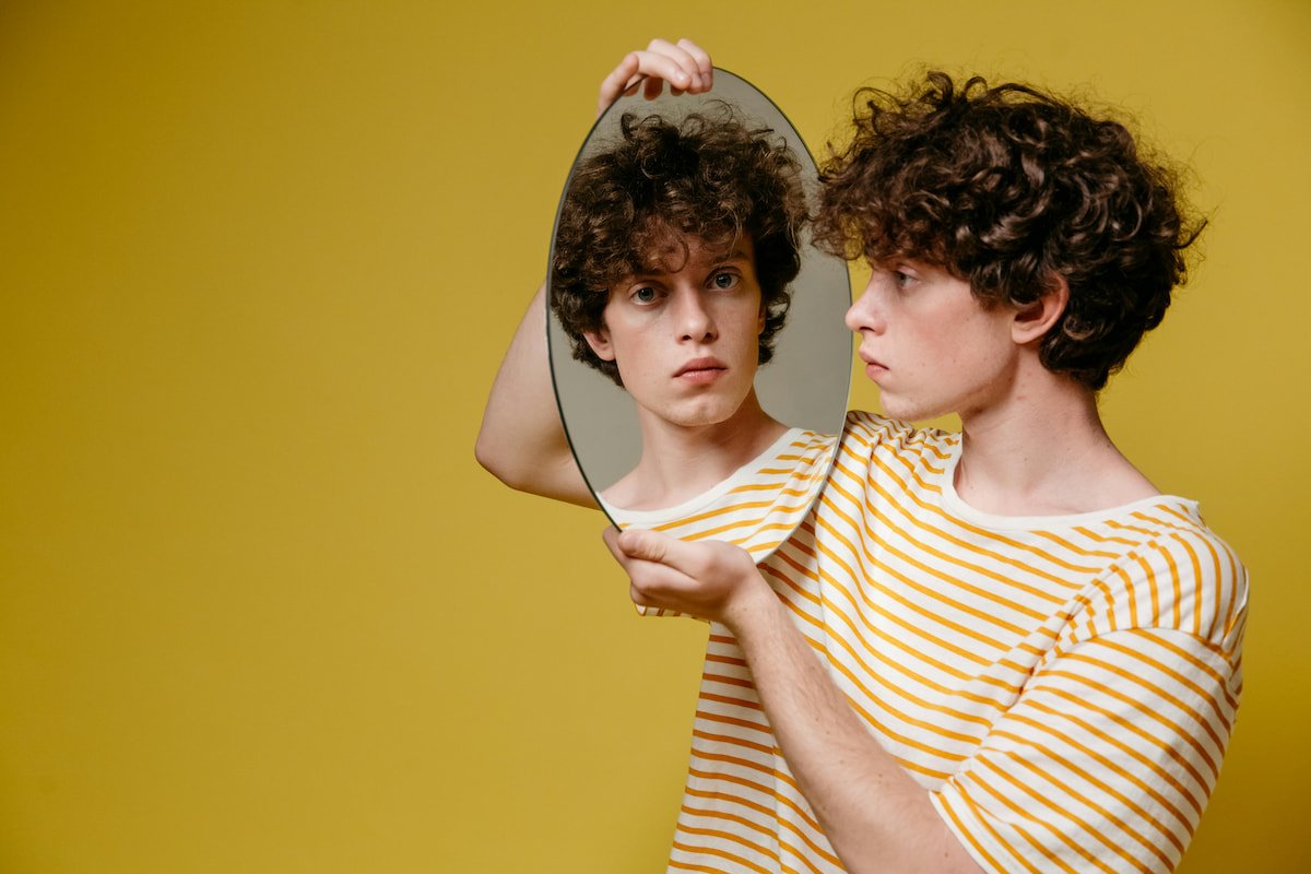 Young man with curly hair looking into a mirror while his reflection looks back, illustrating self-perception and the inner critic