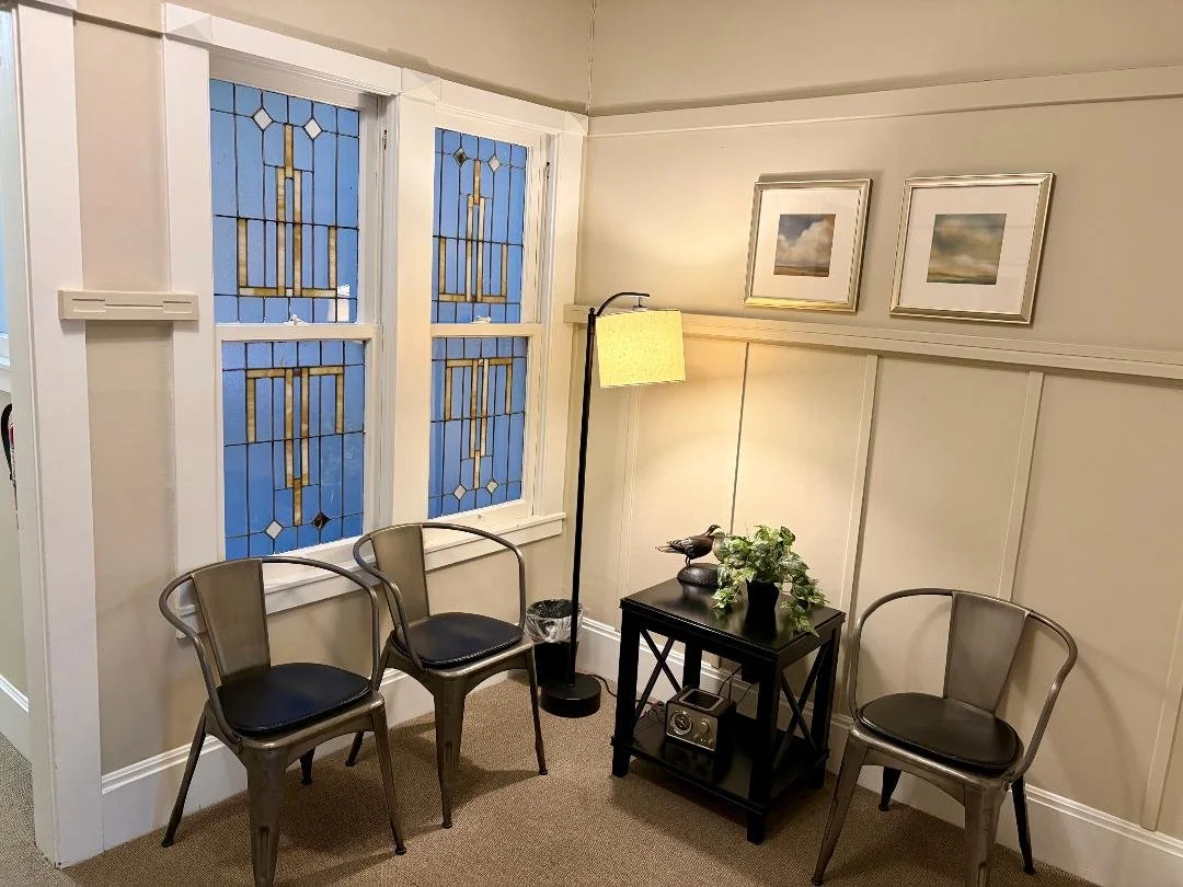 Waiting room with stained glass window, three chairs, floor lamp, side table with plant, framed artwork, and natural light.