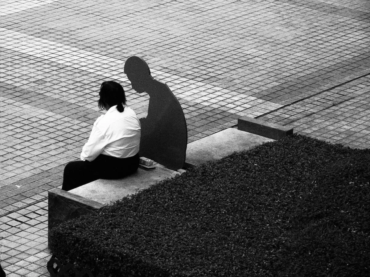 Black-and-white photograph of a woman sitting on a bench in a plaza beside a flat, one-dimensional human figure sculpture.