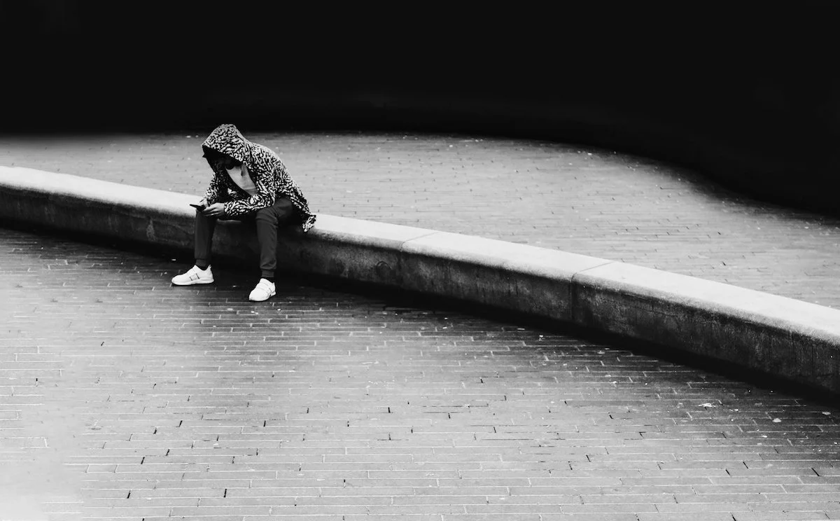 Man wearing a hoodie sitting alone at an empty fountain while looking at his phone.