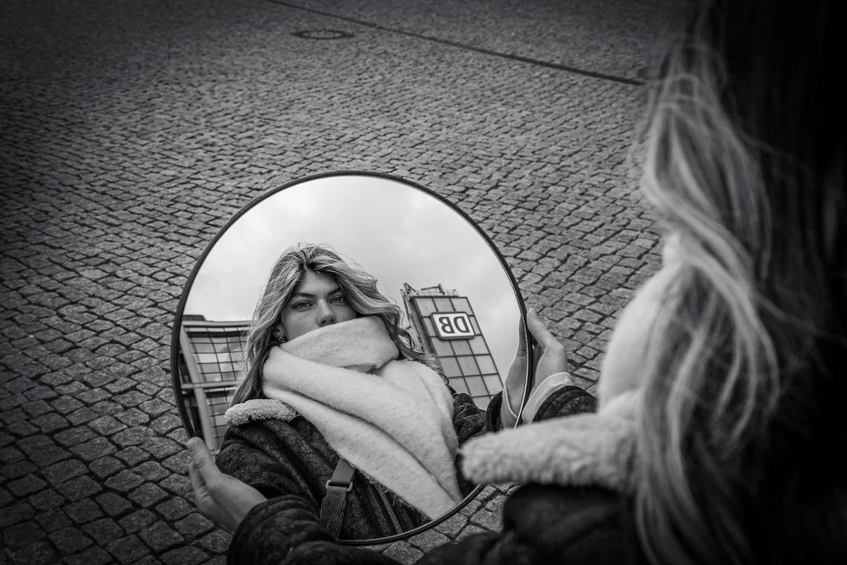 Black-and-white photo of a woman looking at her reflection in a round mirror in an urban setting.
