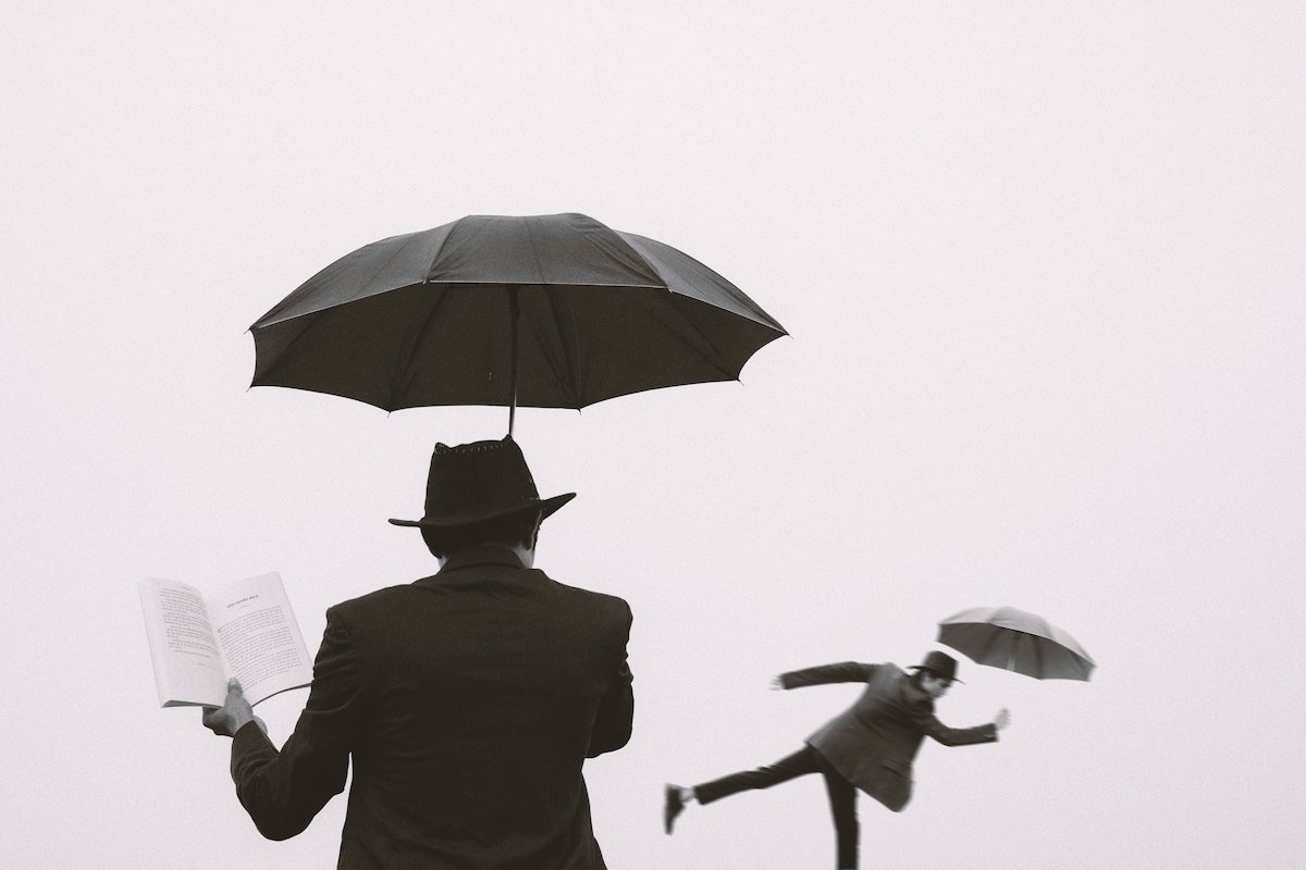 A surreal image of a man reading beneath an umbrella while another version of himself tumbles in the distance.
