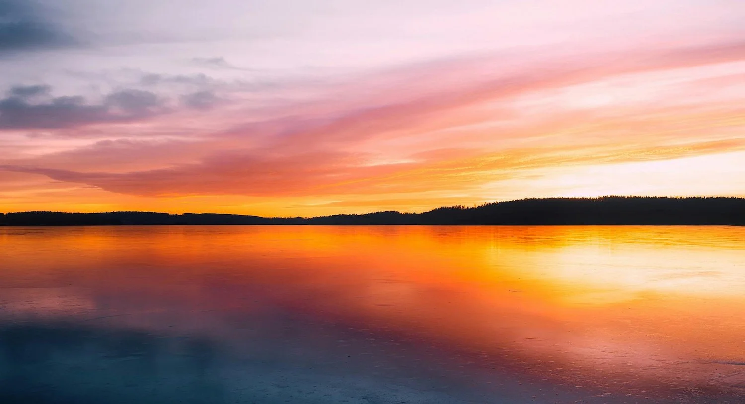 Sunset over a calm lake with colorful pink, orange, and purple sky, silhouetted land in the distance.