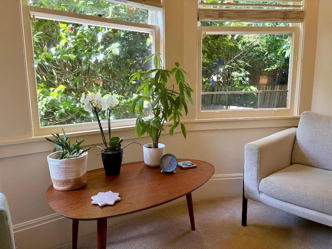 Therapy office with a comfortable chair and mid-century modern table holding an orchid and plants, with bay windows, green foliage, and camellia blossoms in the background.
