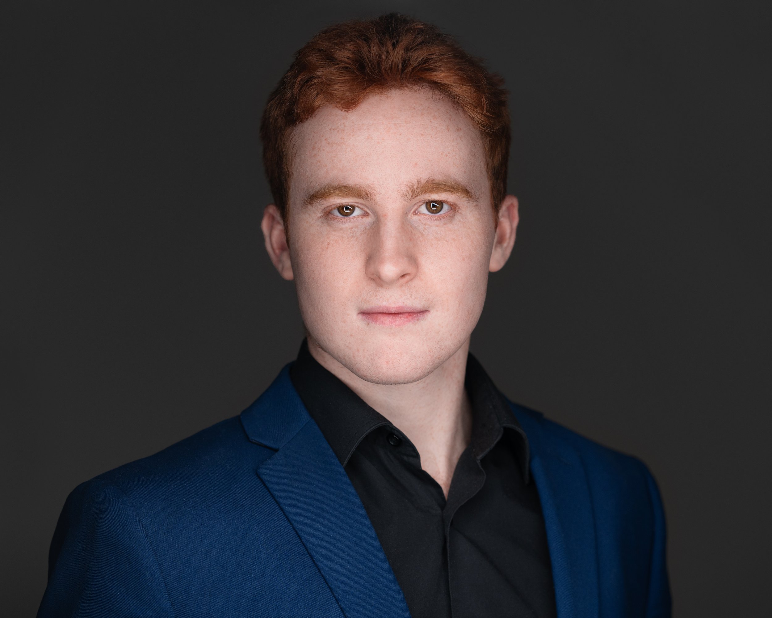 Man wearing a blue suit jacket and black shirt against a gray background.