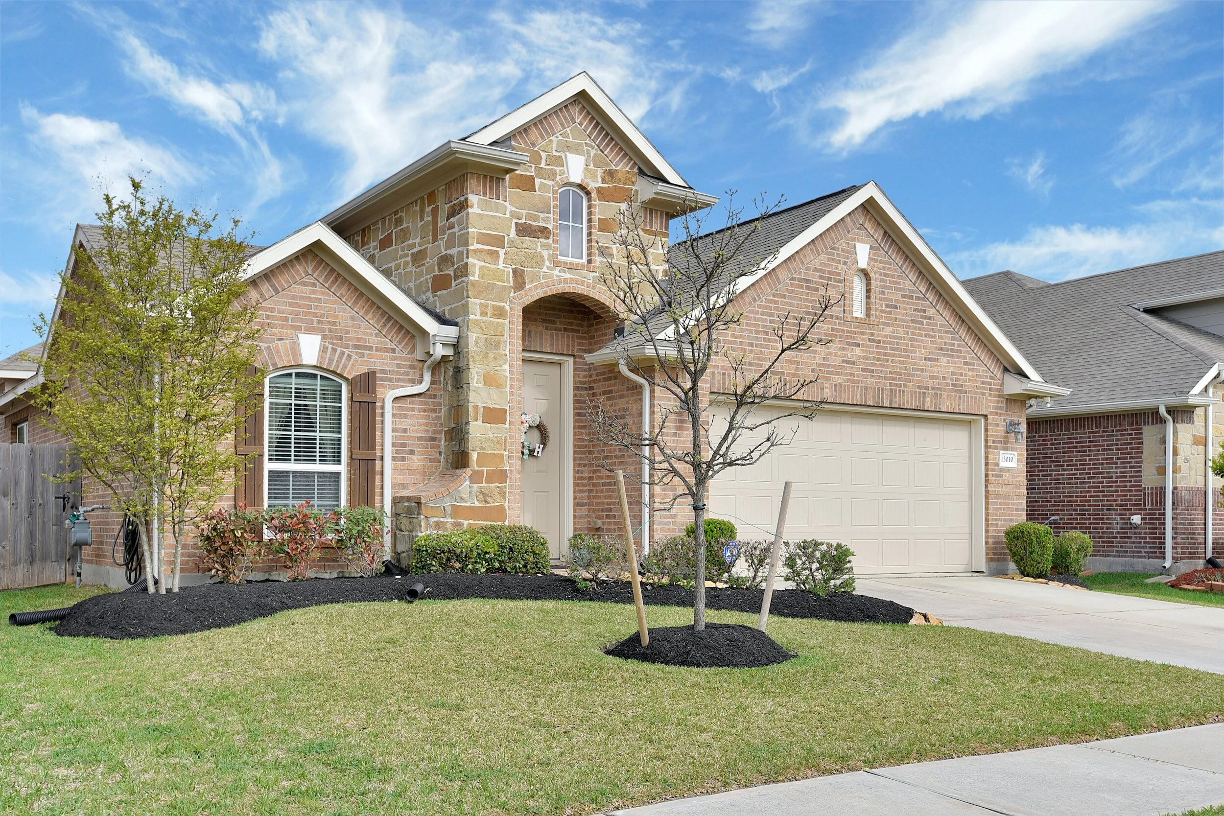 Single-story brick house with attached garage, stone accents, and well-maintained front yard with young trees and shrubs.