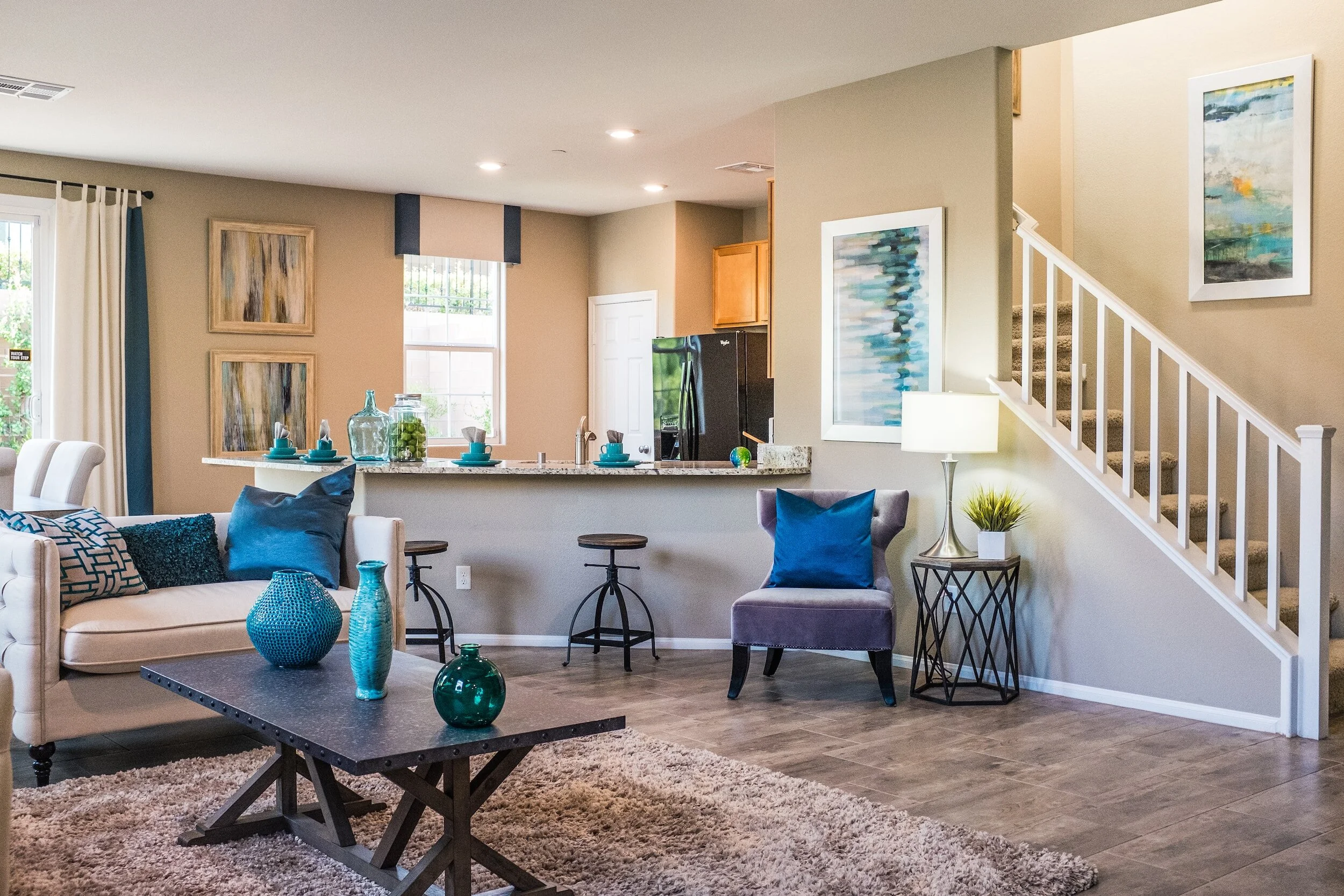 Modern living room with neutral walls and blue accents, including pillows and decorative vases. Features a beige sofa, a gray chair, a wooden coffee table, a shag rug, and a breakfast bar with stools. Artwork and a staircase are visible, creating a cozy, elegant atmosphere.