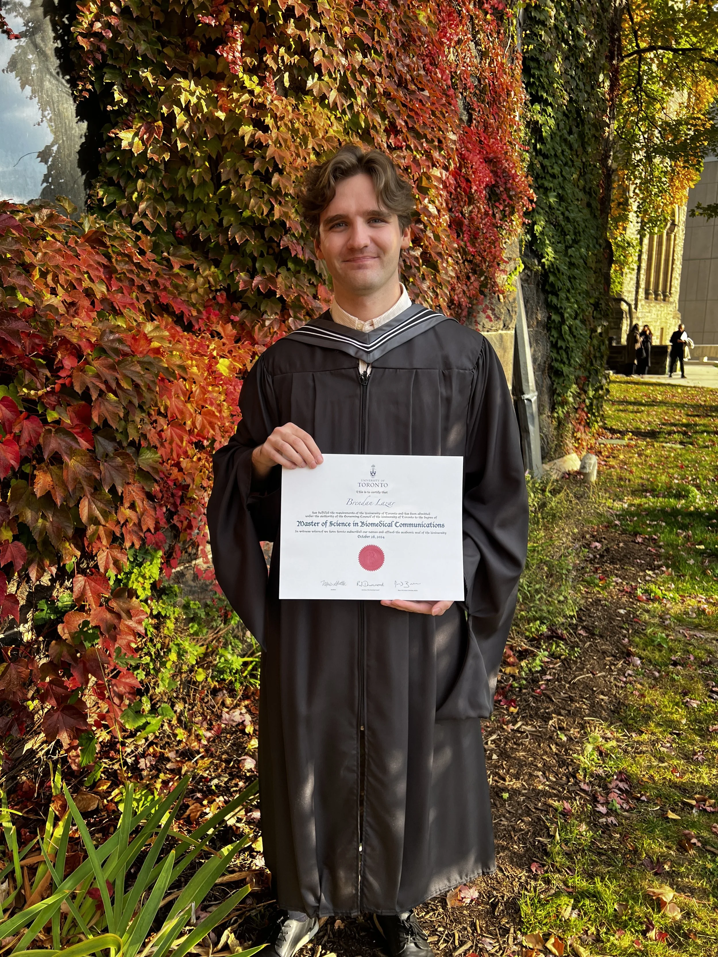 A young man in a graduation cap and gown holding a diploma stands outdoors among autumn-colored leaves and foliage, smiling at the camera.