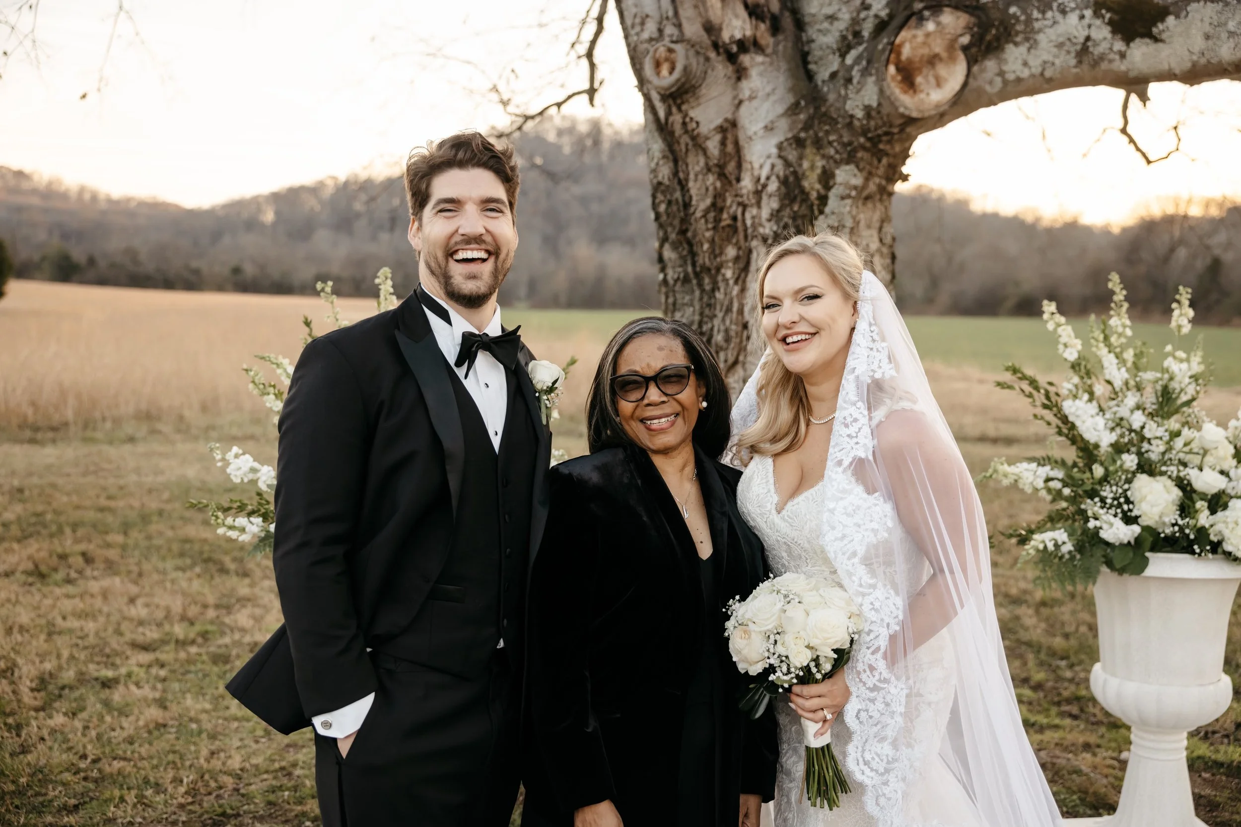 Rev. Benita in her elegant black suit stands in between the groom who wears a black tuxedo and the bride who wears a spaghetti strap dress with a long lace veil under a tree at Southall Meadows Farm and In in Franklin, TN.