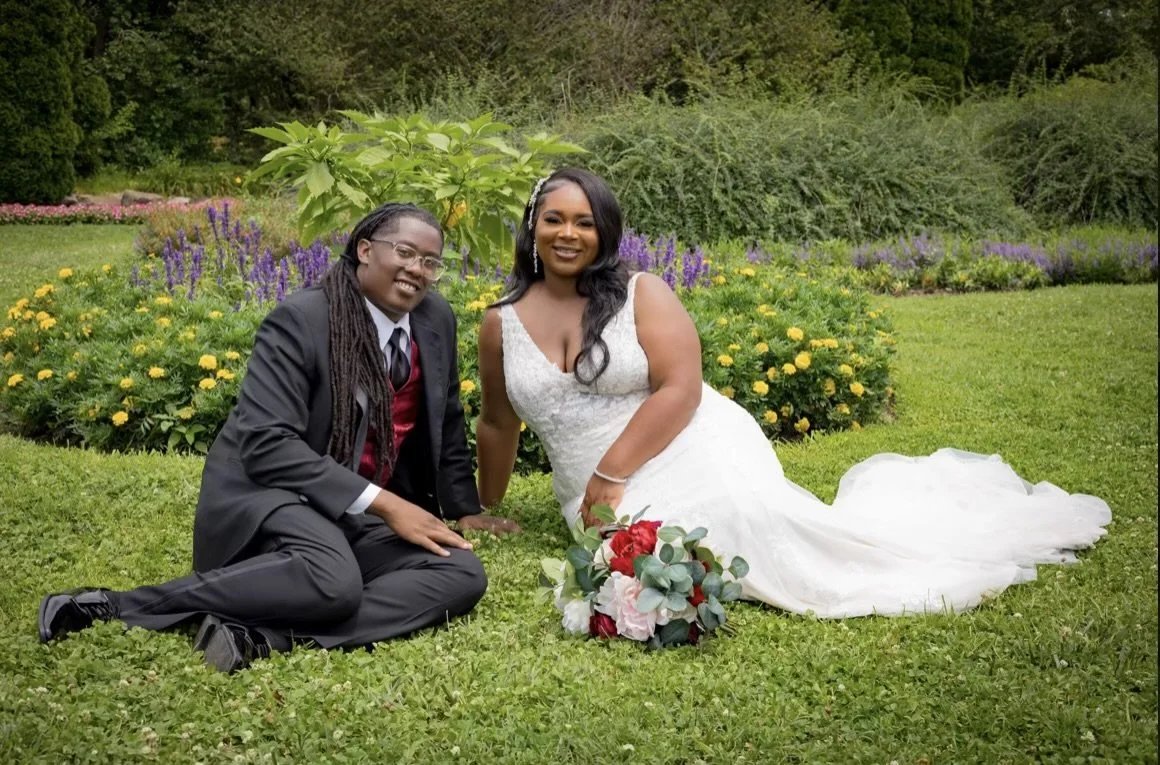 The groom, wearing a black suit with red vest and the bride wear a v-cut lace wedding gown is sitting on the beautiful floral grounds at Centennial Park in Nashville, TN.