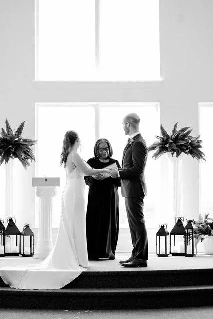 At New Hope Chapel in Nashville, TN the bride wearinfg a flowinf A-line gown and the groom wearing a black tuxedo hold hands together while intently listening to Rev. Benita as she officiates their wedding.