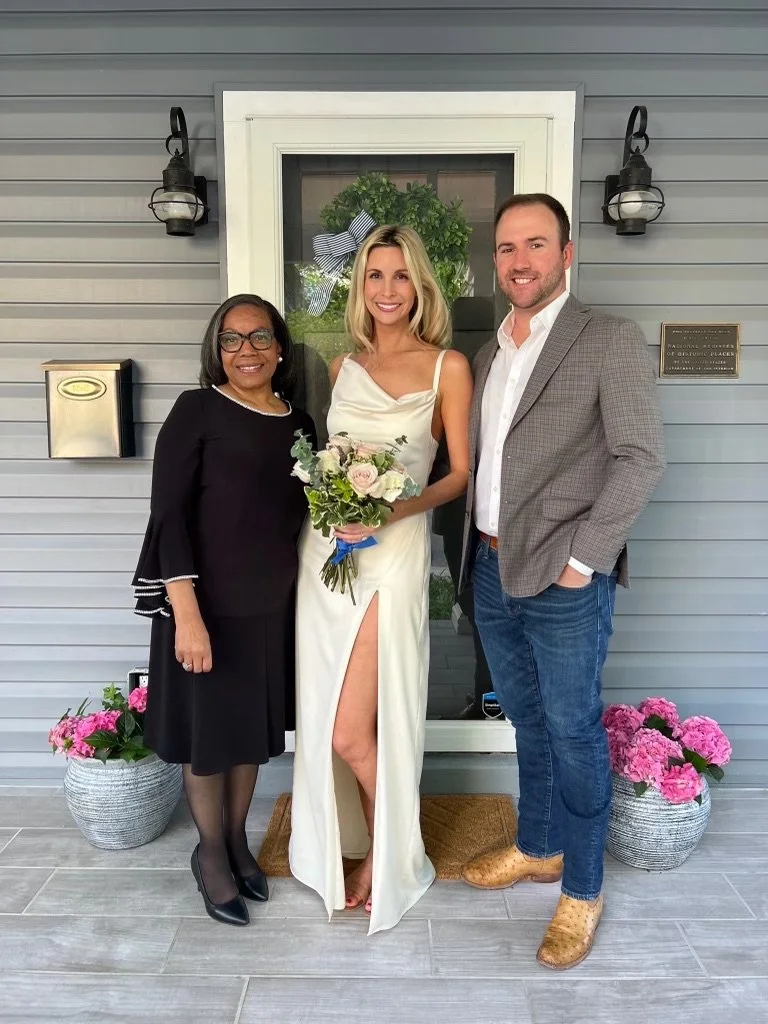 Rev. Benita stands next to the bride, who is wearing a vintage gown, while the groom, dressed in a casual blazer and denim jeans, poses for a picture alongside them.