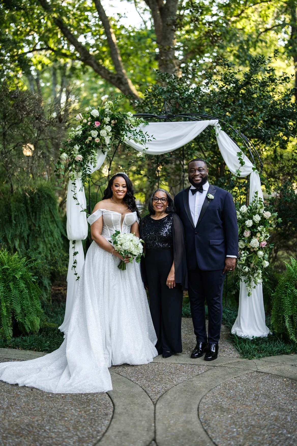 At the Riverwood Mansion in Nashville, TN, the bride, in an off-the-shoulder princess-style gown, and the groom, dressed in a navy blue tuxedo, stand beside Rev. Benita under a white sheer draped archway..