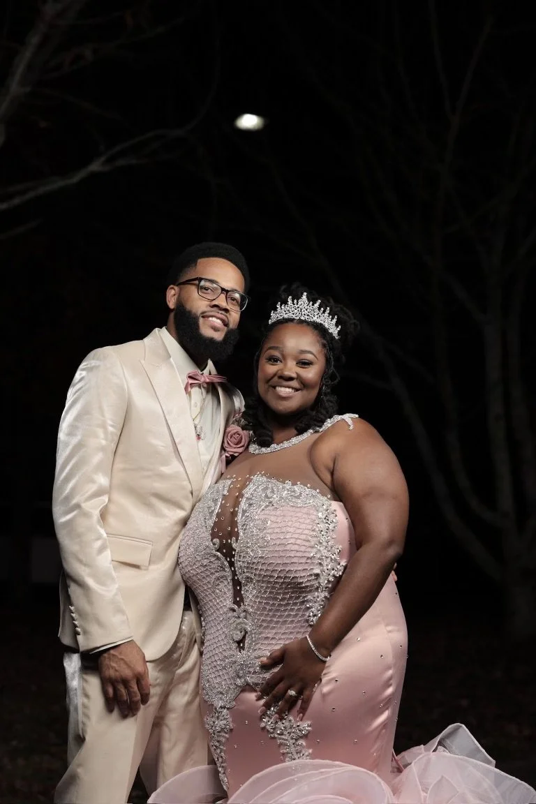 The groom is standing near hi bride in an off white shimmering suit with rose gold accents , stands beside hi bride who wears a lace rose gold gown highlighted with a diamond embellished crown.