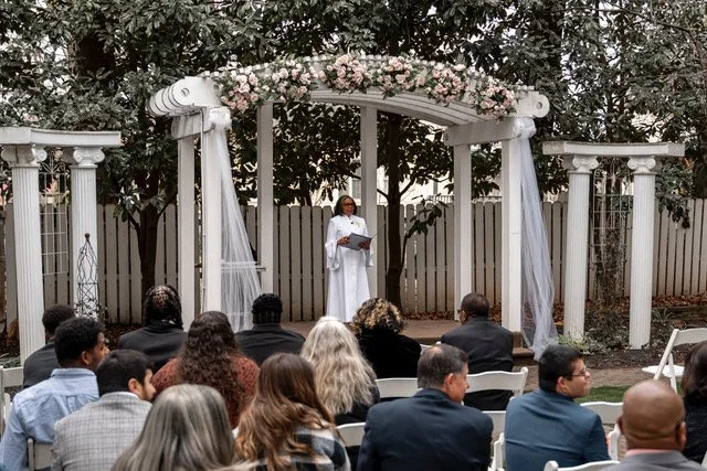Rev. Benita is standing before the attendees under a beautifully floral draped arch awaiting the wedding party.
