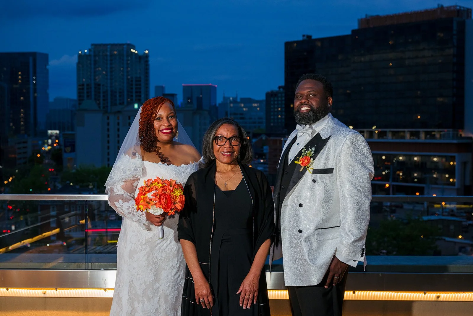 Standing on the balcony overlooking the city lights at the Kimpton Aeration Hotel the bride wearing an off-the-shoulder gown with a citrus colored bouquet and the groom wearing a silver and black tuxedo stand beside the officiant, Rev. Benita.