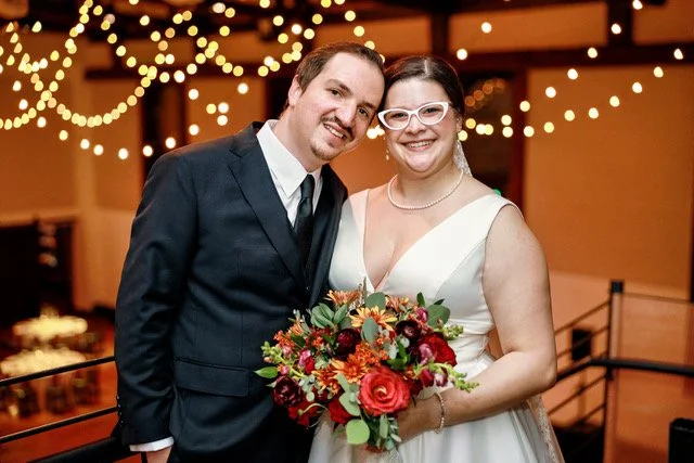 Bride in a white dress holding a bouquet sits next to groom in a black suit on a leather couch against a brick wall. Both smiling.