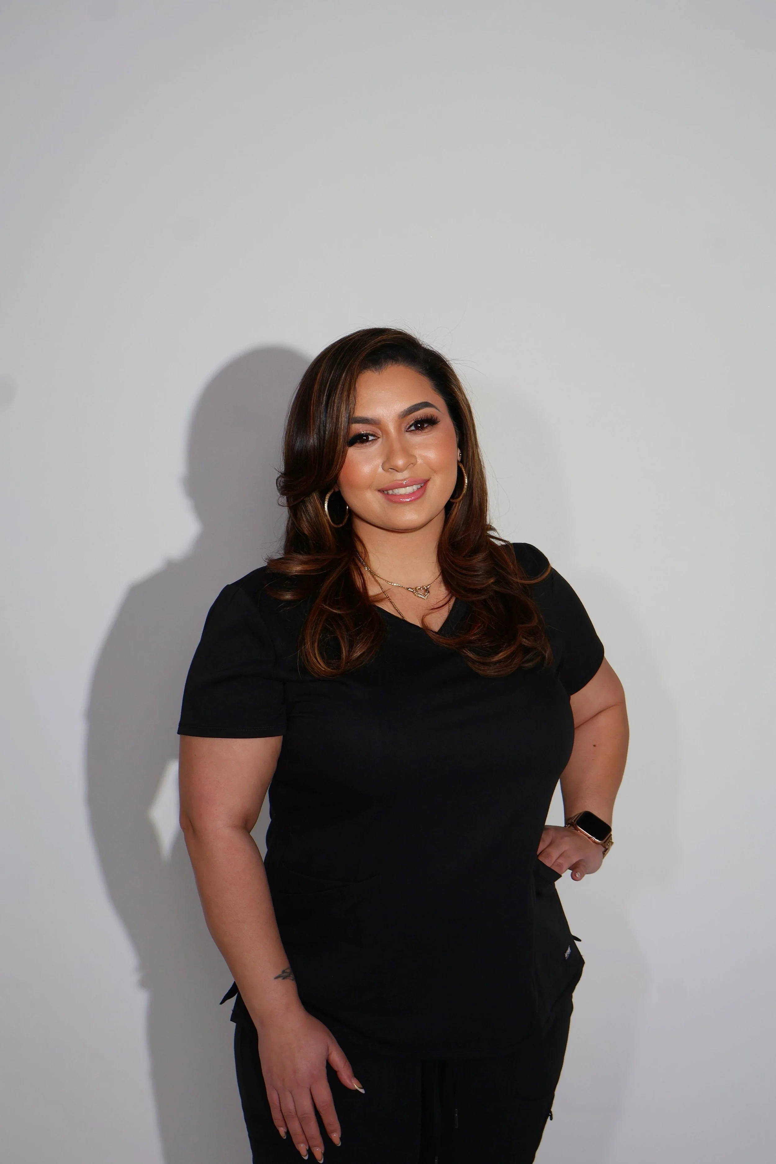 A woman with long wavy brown hair, wearing a black t-shirt and black cargo pants, standing against a plain white wall. She is smiling and has gold hoop earrings, a layered necklace, and a smartwatch on her left wrist.