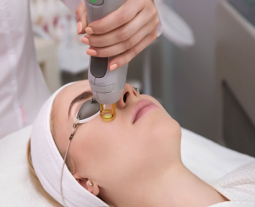 A woman undergoing a facial treatment with a handheld device emitting a yellow light, in a clinical setting.