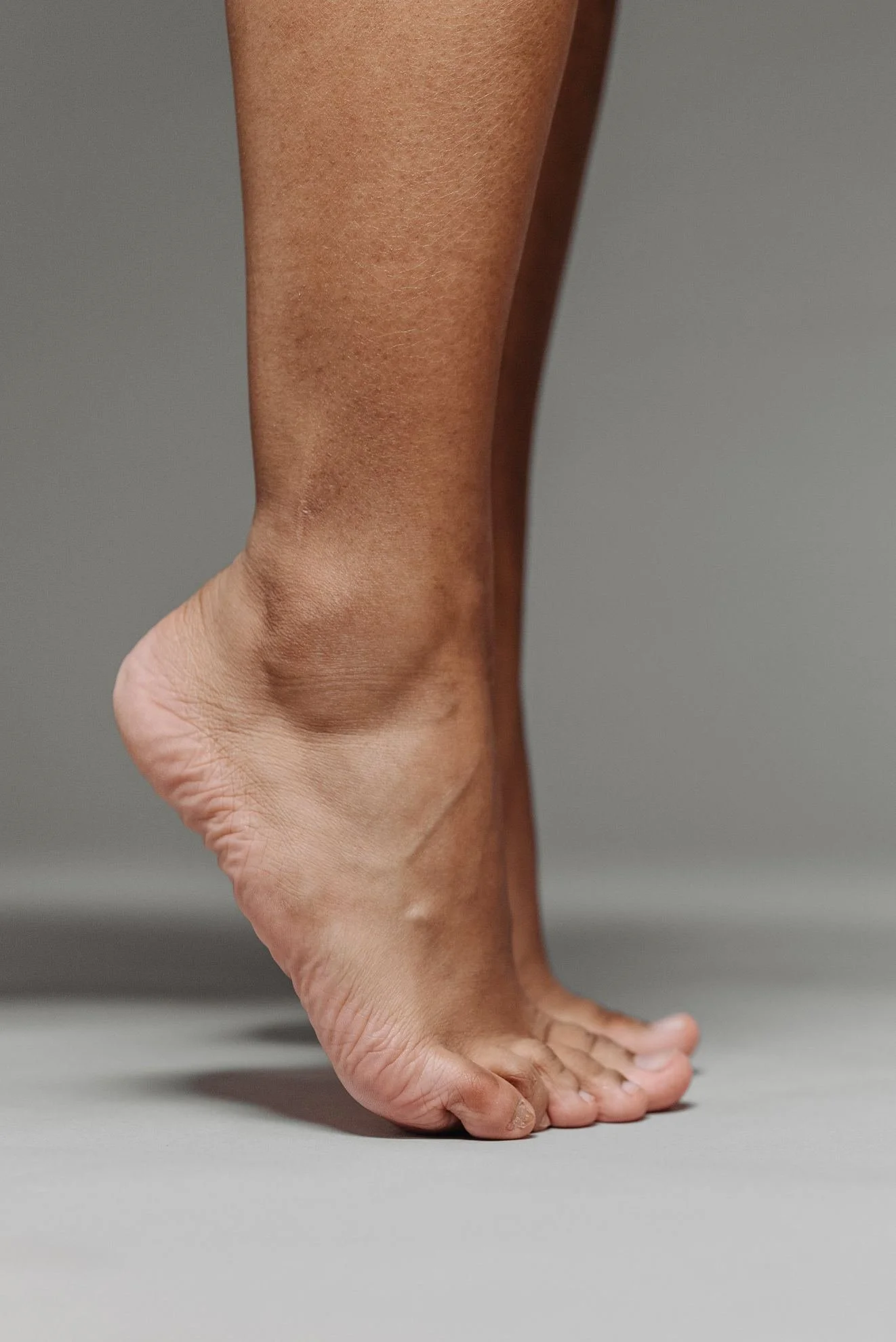 A person poses in a handstand with one hand on the ground and the other arm extended upward, balancing on the side of their face, against a plain gray background.