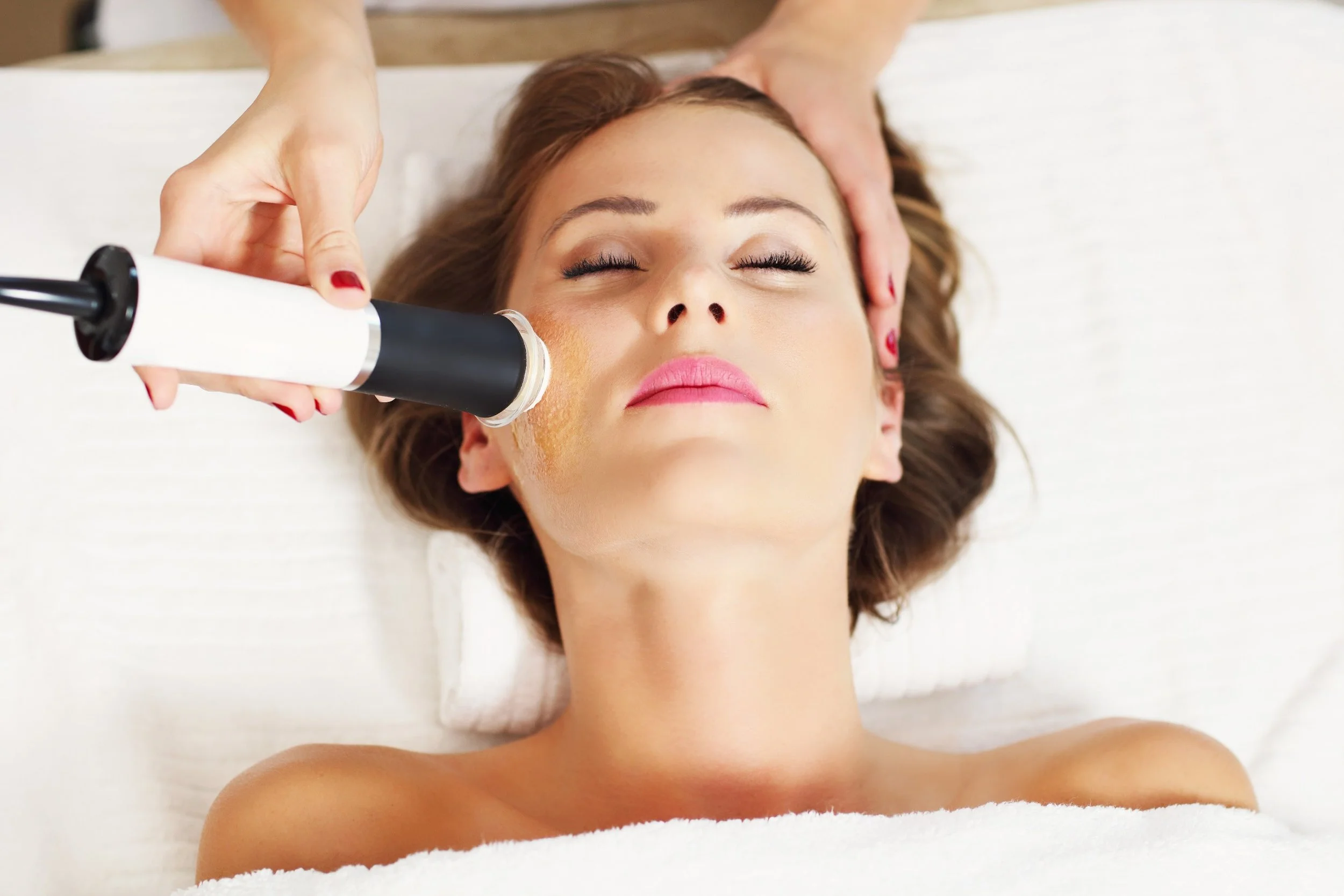 A woman lying on a treatment bed with her eyes closed, receiving a facial massage or skin treatment using a handheld device.