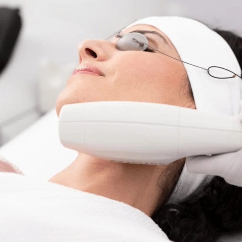 A woman undergoing a facial treatment in a spa or clinic, wearing protective eye goggles and with a skincare device on her face.