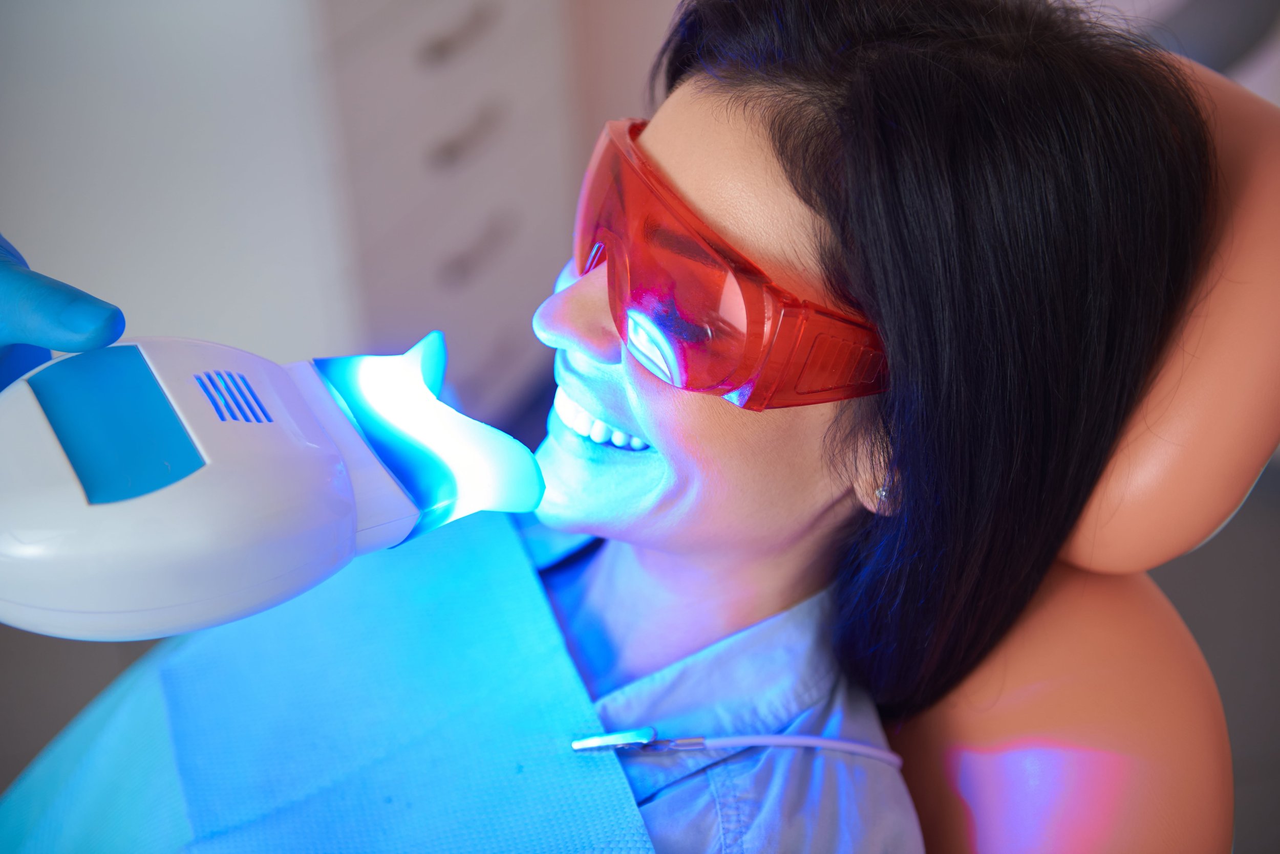 A woman with dark hair wearing red protective goggles getting a teeth cleaning with blue light from a dental device.