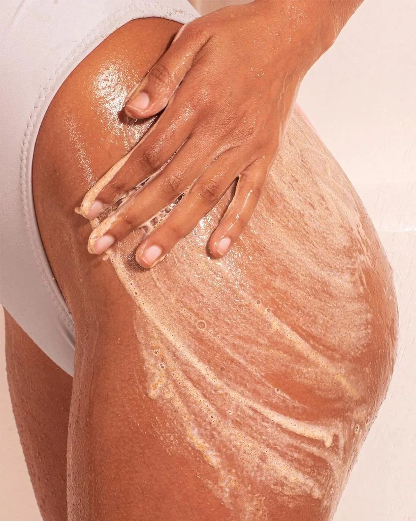 Person applying exfoliating scrub to their thigh in a bathroom or spa setting.