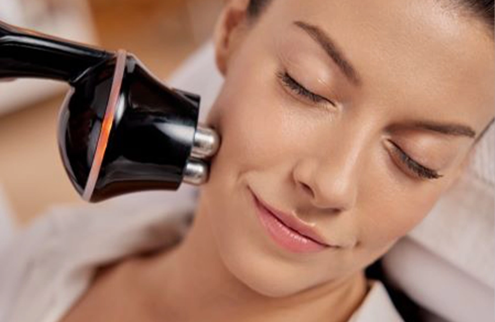 A woman receiving a facial treatment with a black facial device applied to her cheek.