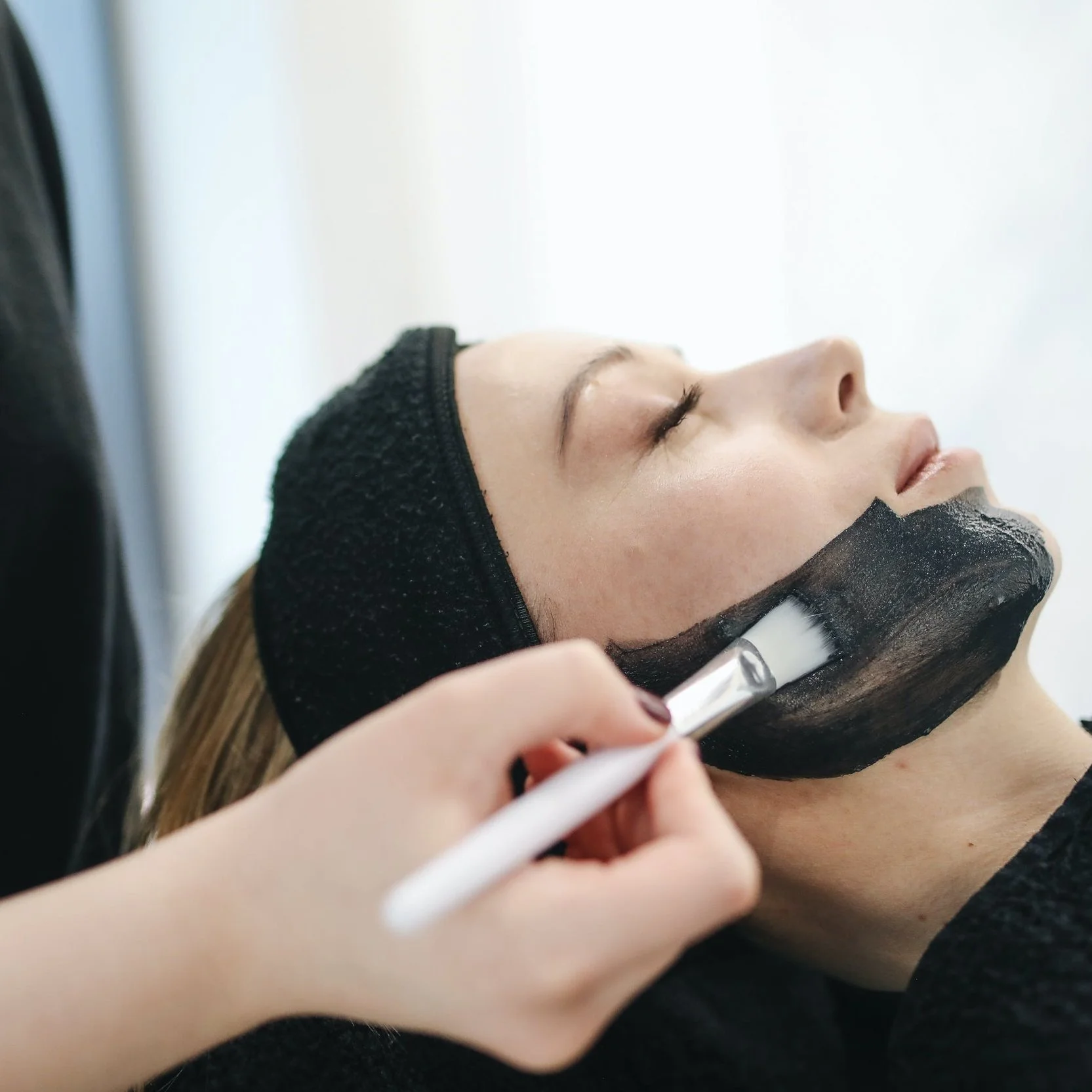 A woman lying on her back receiving a facial treatment, with a black headband and mask, as an esthetician applies a facial mask with a brush.