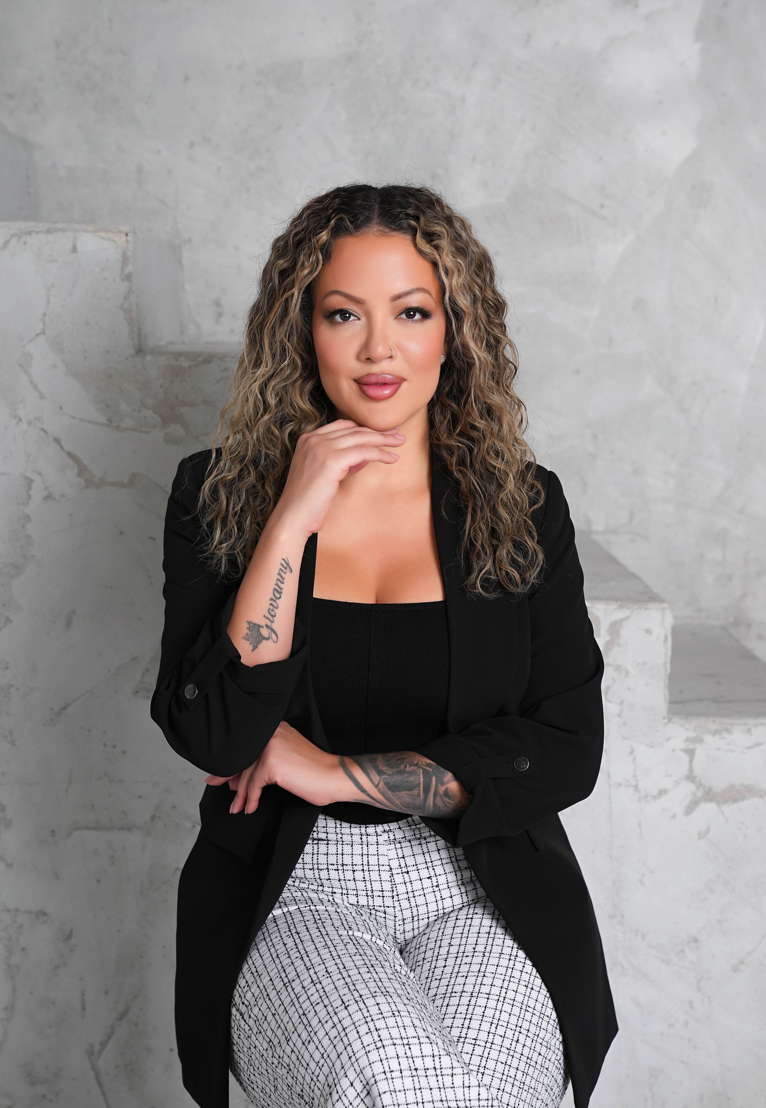 A woman with curly hair and tattoos on her arm is sitting in front of a gray textured wall, wearing a black blazer, black top, and patterned pants. She has makeup on and is looking directly at the camera with one hand resting under her chin.