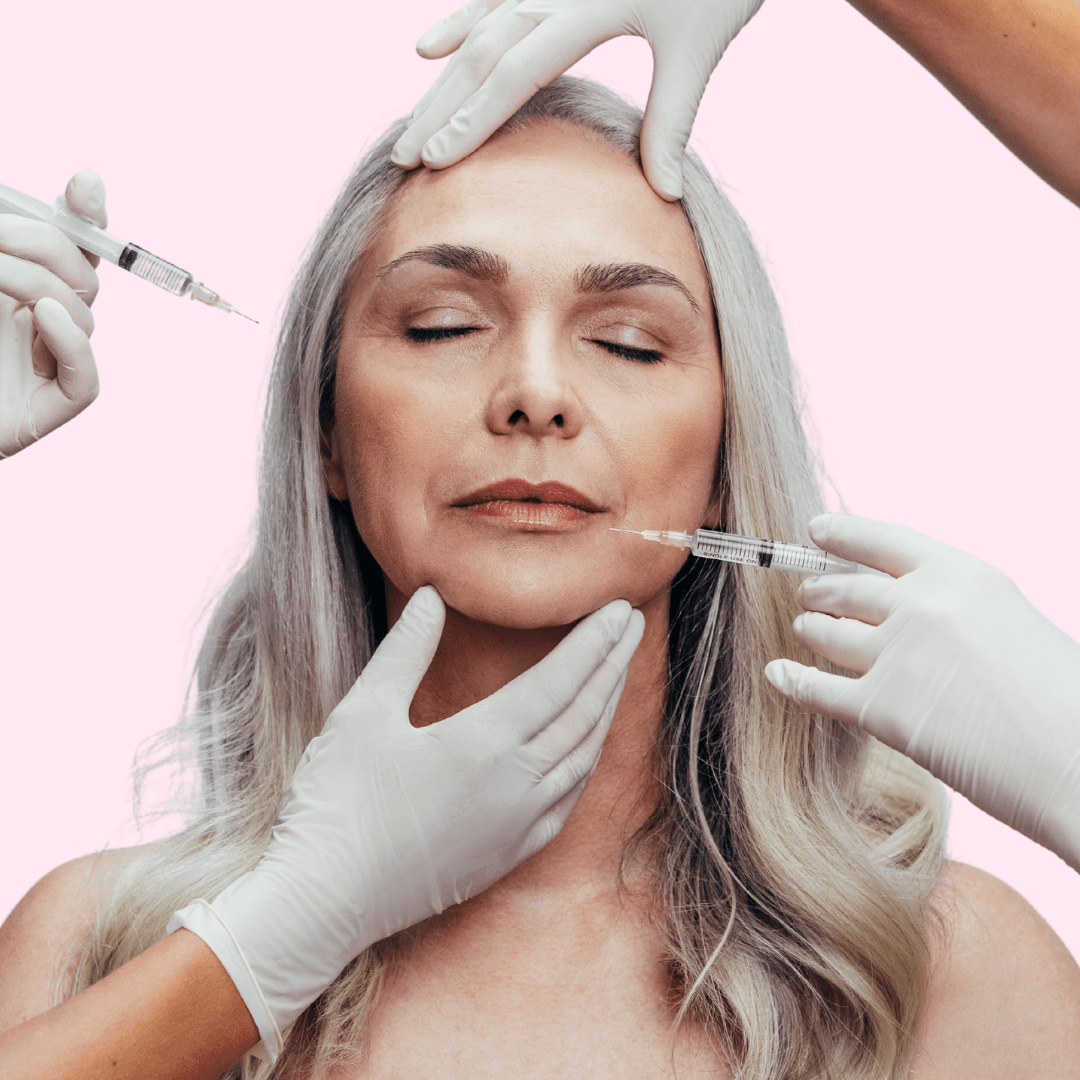 A mature woman with silver hair receiving cosmetic injections in her face from medical professionals wearing white gloves, against a pink background.