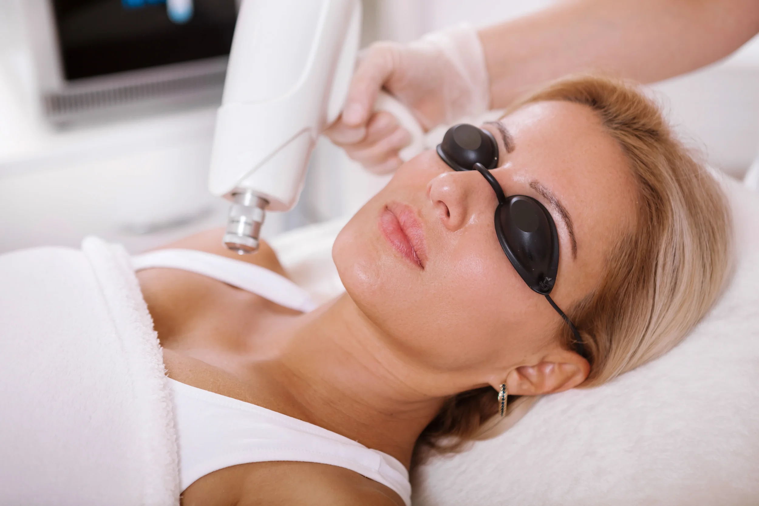 A woman with blonde hair and earrings lying on a treatment bed. She is wearing protective goggles and receiving a facial treatment with a handheld device.
