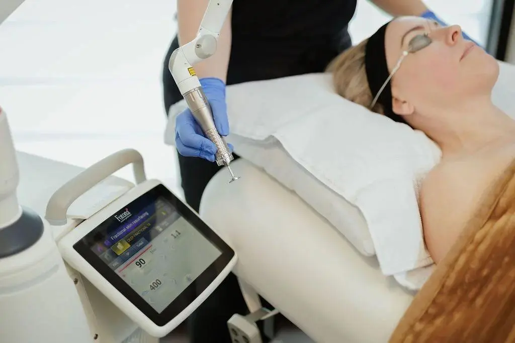 A woman undergoing a facial treatment with a handheld device emitting a yellow light, in a clinical setting.